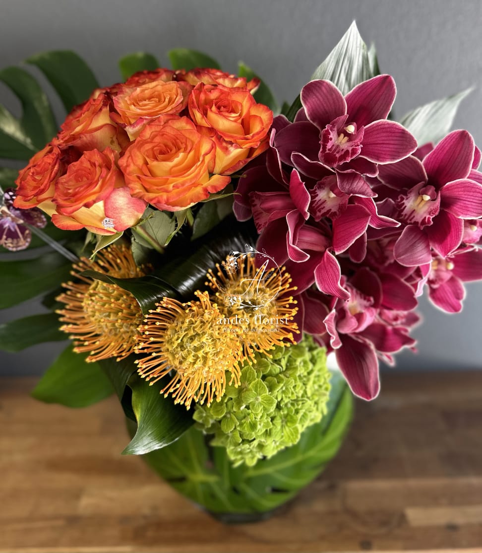 Trifecta - Orchids Roses and more - If you're looking for something modern, unique, and long-lasting, look no further! This perfect Trifecta is a colorful abundance of roses, orchids, and tropical florals. In a stunning vase with tropical leaf accents, this design is SUCH a vibe. 