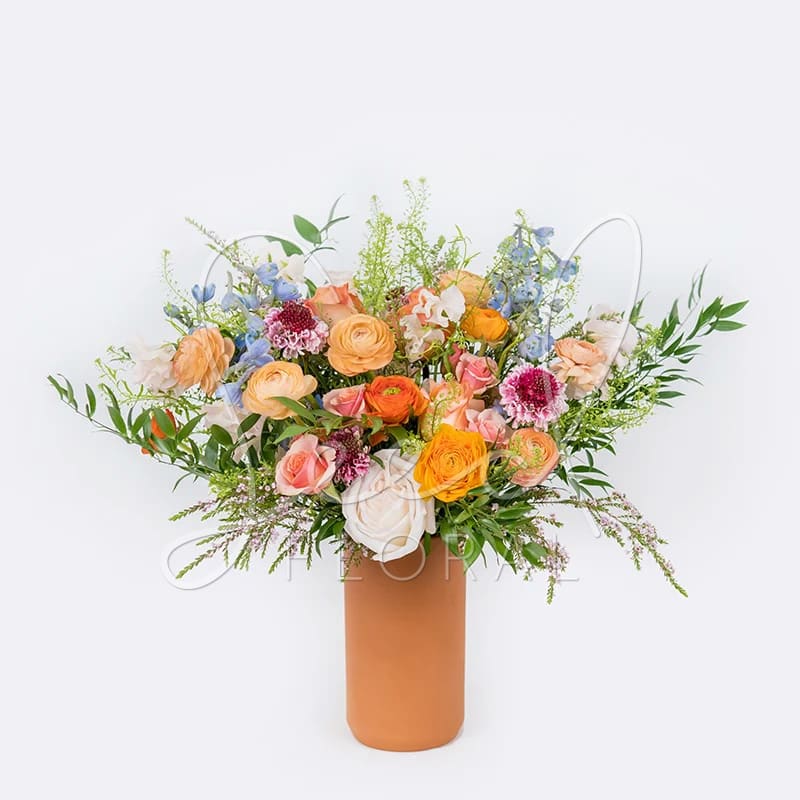Sunnier Skies - Find your happy place with this vibrant tall design bursting with colorful seasonal blooms  in shades of peach, orange, and blue designed with foliage inside our Kendal Terra-Cotta vase 8&quot; x 4&quot;.  (Arrangement is one sided)
