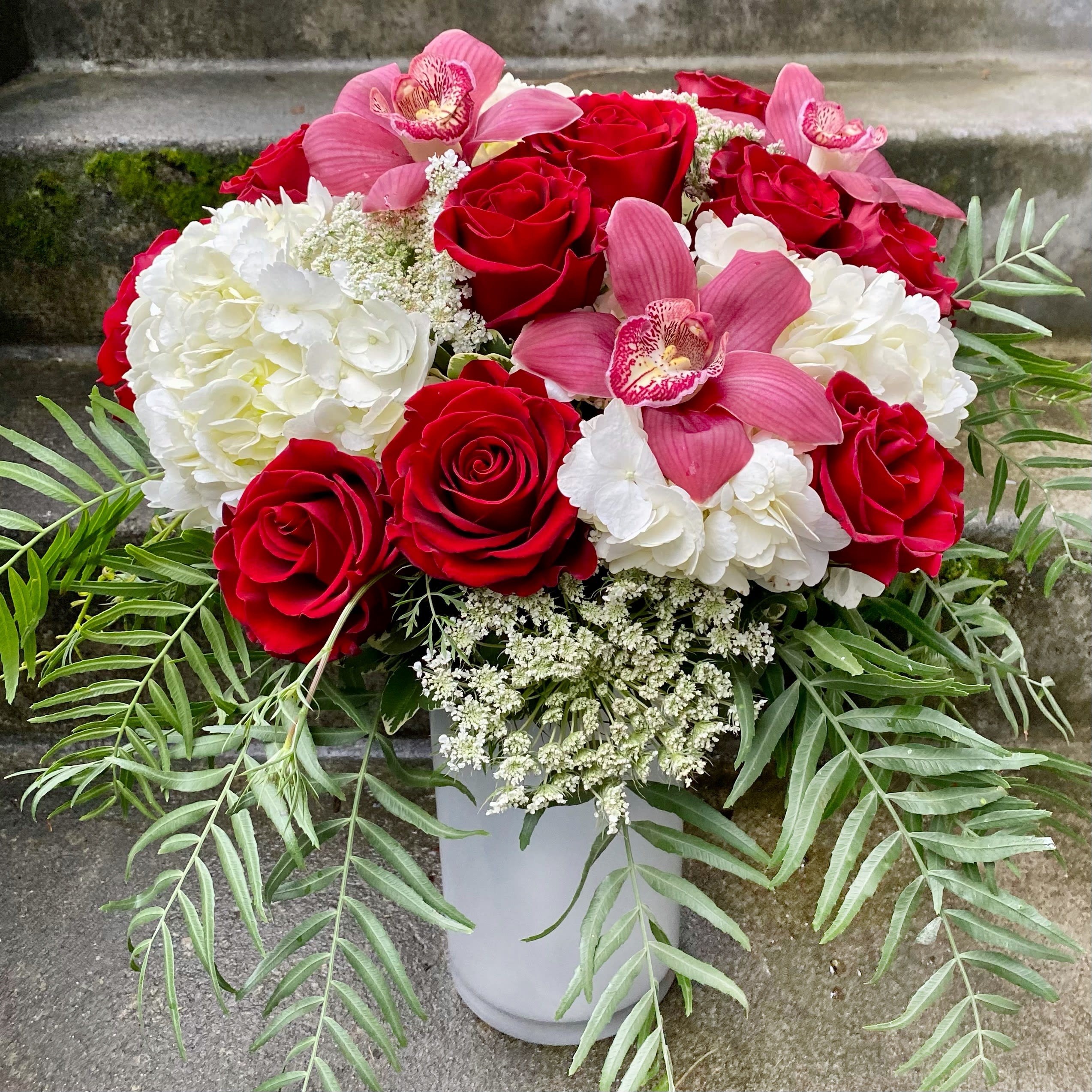 One Dozen Signature Red Roses - Our Signature Rose Arrangements are the epitome of our design style. Rather than focusing on stem length and filler, our signature roses have one-dozen perfect red roses tucked into pillows of fluffy hydrangeas accented with seasonal flowers and foliage. Cymbidium orchids serve as the perfect finishing touch, giving all our signature rose arrangements that special Fiori flair! The result: an arrangement that is beautiful, lush and just a little bit decadent.  The vase is a 10-inch glass cylinder, whose clean lines complement the arrangement while allowing the focus to stay on the flowers. The vase is available in white or in a clear glass lined with synthetic variegated ti leaves that make the roses really pop! We also offer our Signature Roses in a versatile low cylinder vase. If you prefer the lower style, let us know by leaving a note in the “Special Instructions” section during checkout.  The Deluxe version includes even more accent flowers and orchids, while the Premium option upgrades the arrangement to eighteen red roses in a 12-inch vase.  Approximate dimensions - Standard and Deluxe: 14 inches wide and 17 inches tall. Premium: 15 inches wide and 20 inches tall.  Orders of this Fiori Floral Design original arrangement for delivery to an address outside of our standard delivery area will likely require substitutions of the vase, accents or greenery by the local florist who fills your order. You can check whether a recipient’s address is in our standard delivery area before you order by looking at the list of Neighborhoods, Cities and Zip Codes at the bottom of our home page or by giving us a call at (206) 329-3944.