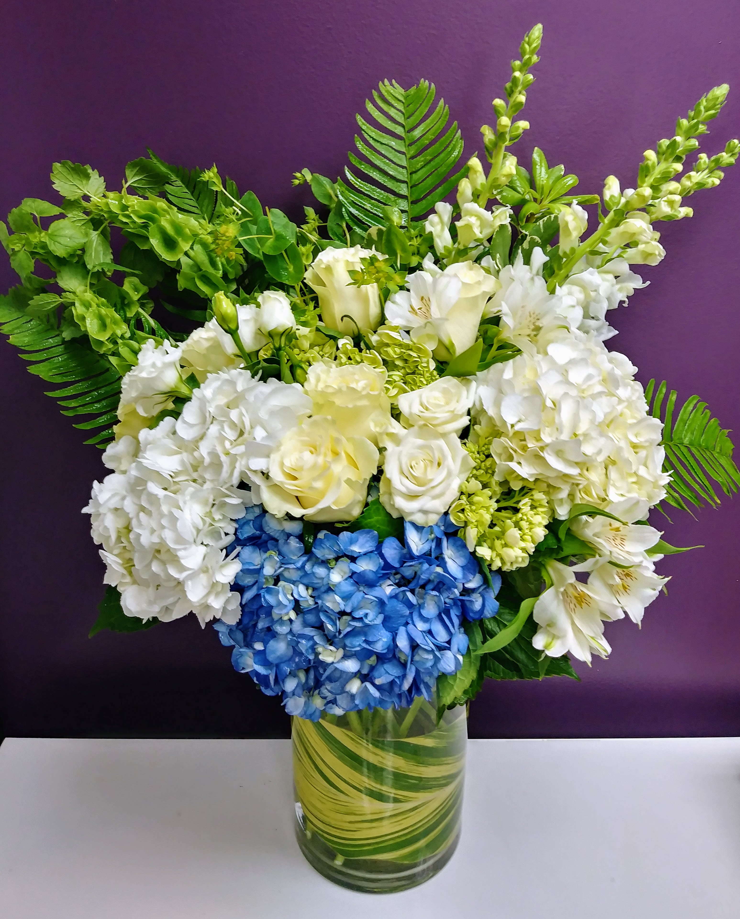  Ely's - Fill any day with the beauty of the European countryside . A sweet combination of Blue, White and Green Hydrangeas, White and cream Roses, White Lisianthus, Bells of Ireland and ferns. 