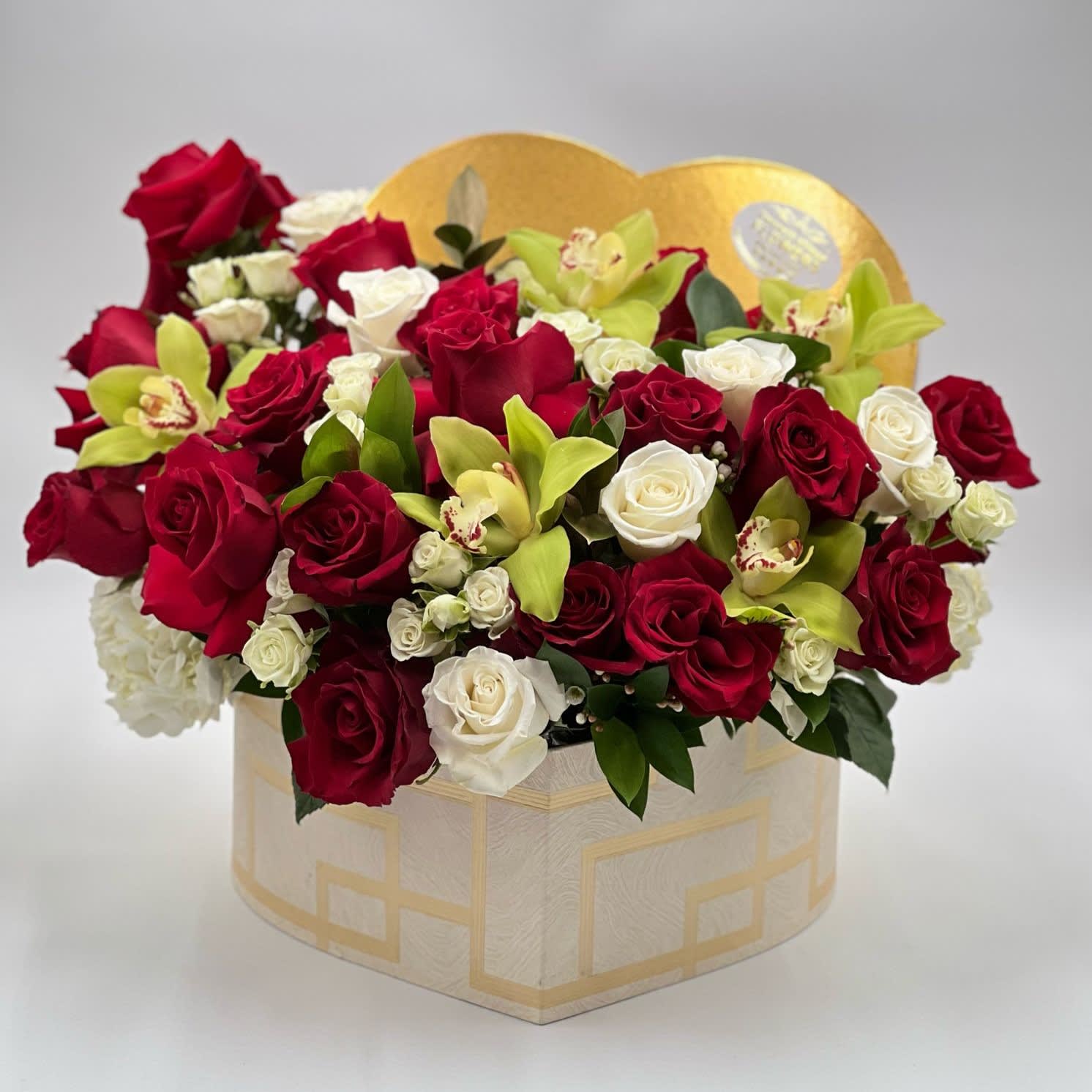 Love Box - This luxurious floral arrangement nestled in an elegant, cream-colored octagonal box with geometric patterns. The arrangement highlights blend of red and white roses, interspersed with delicate greenery and exotic green orchids, adding a touch of sophisticated variety. The backdrop is a golden heart, which complements the rich colors of the flowers, suggesting that this could be a premium gift for a Valentine's Day or an anniversary.