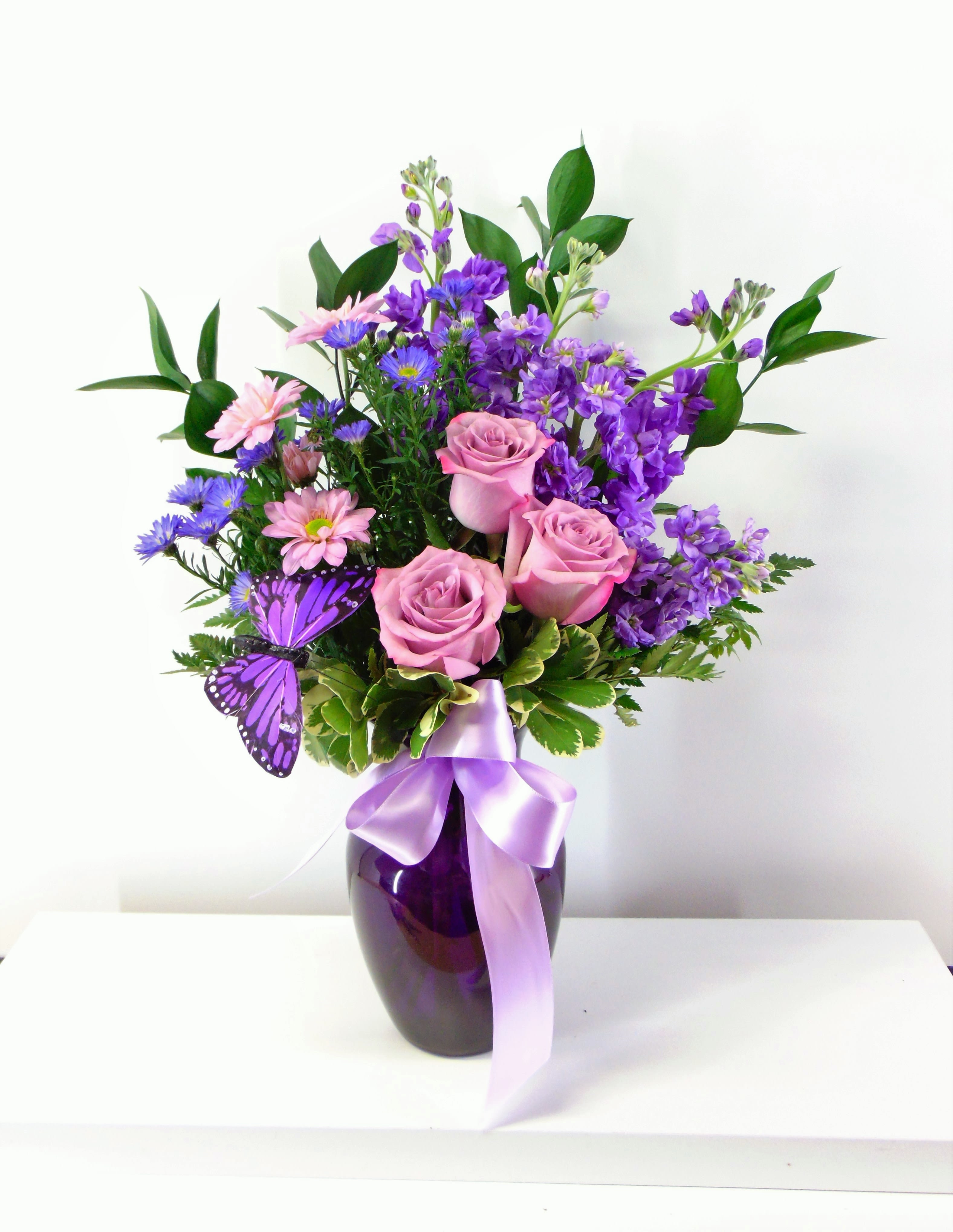 Violet Visions - Purple stock, purple asters, lavender daisies and lavender roses in a purple vase with satin ribbon purple butterfly. The standard option is pictured.  The deluxe option adds 3 more stems. The premium option is larger and includes 6 more stems of flowers.  All sizes include the satin bow and butterfly.