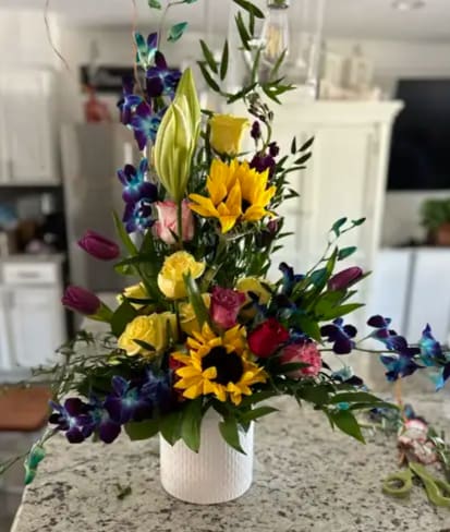 Vivid Cascade Vivid Cascade - The bouquet is a lively collection of colors and shapes, featuring sunny yellow sunflowers as a cheerful focal point. These are complemented by deep purple tulips and rich blue delphiniums, which add a sense of drama and height to the arrangement.