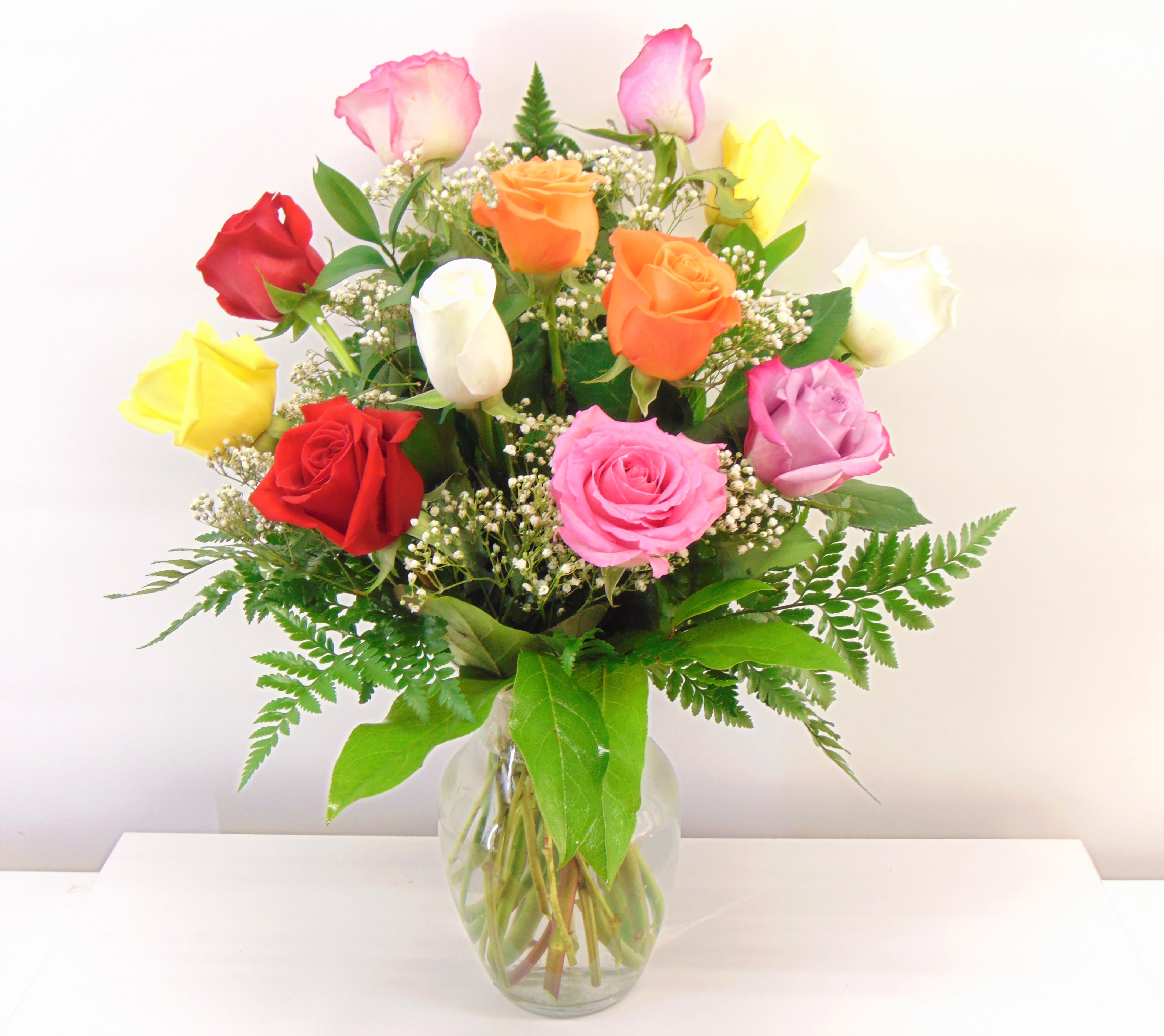 Rainbow Roses - One dozen assorted color roses arranged with baby's breath and greenery in a clear glass vase. 