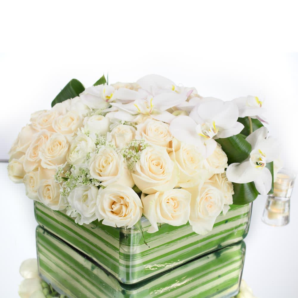 Pure White - During this time of mourning, our funeral flower arrangement offers a heartfelt tribute that honors your loved one's memory with grace and beauty. This exquisite arrangement includes a selection of white roses, white Phalaenopsis blooms, white ranunculus, and filler greens, expertly arranged in a square glass vase. To add additional accents and details, we incorporate thoughtful elements that enhance the overall aesthetic and symbolism of the arrangement.  The white roses, symbolic of love, reverence, and purity, are elegantly arranged as the focal point of this tribute. Their timeless beauty and delicate petals serve as a representation of eternal remembrance and honor for your departed loved one.  Complementing the roses, the white Phalaenopsis blooms add an ethereal touch to the arrangement. These graceful orchids, known as &quot;Moth Orchids,&quot; symbolize elegance and hope, serving as a gentle reminder of the beauty and resilience of life. Their slender stems and exquisite flowers bring a sense of serenity and calmness to the arrangement.  The arrangement is further adorned with white ranunculus, which adds layers of delicate petals and a touch of charm and beauty. The ranunculus flowers represent a graceful farewell, offering solace and comfort during this time of loss.  To enhance the overall visual appeal, filler greens are carefully selected and incorporated into the arrangement. These fresh greens contribute depth and texture, providing a lush backdrop that represents life and growth. This adds a sense of vibrancy and natural harmony to the tribute.  With the square glass vase, the arrangement receives a modern and sophisticated touch. The transparent nature of the vase allows the beauty of the flowers to be showcased fully, while the clean lines and geometric shape add a contemporary element to the arrangement.  To add additional accents and details, our skilled florists include thoughtful elements such as delicate ribbons, subtle pearls, or small clusters of baby's breath. These details are chosen to enhance the overall visual impact and provide a personalized and meaningful touch.  With each meticulously arranged element in this funeral flower arrangement, we hope to create a poignant and beautiful tribute that honors your loved one's life, expresses your heartfelt condolences, and provides a source of comfort and peace during this difficult time.  Please rest assured that our professional florists select only the freshest blooms and arrange them with precision and care, ensuring that this arrangement reflects the utmost respect and sensitivity.  May this funeral flower arrangement serve as a lasting symbol of love, remembrance, and support for all those affected by the loss.