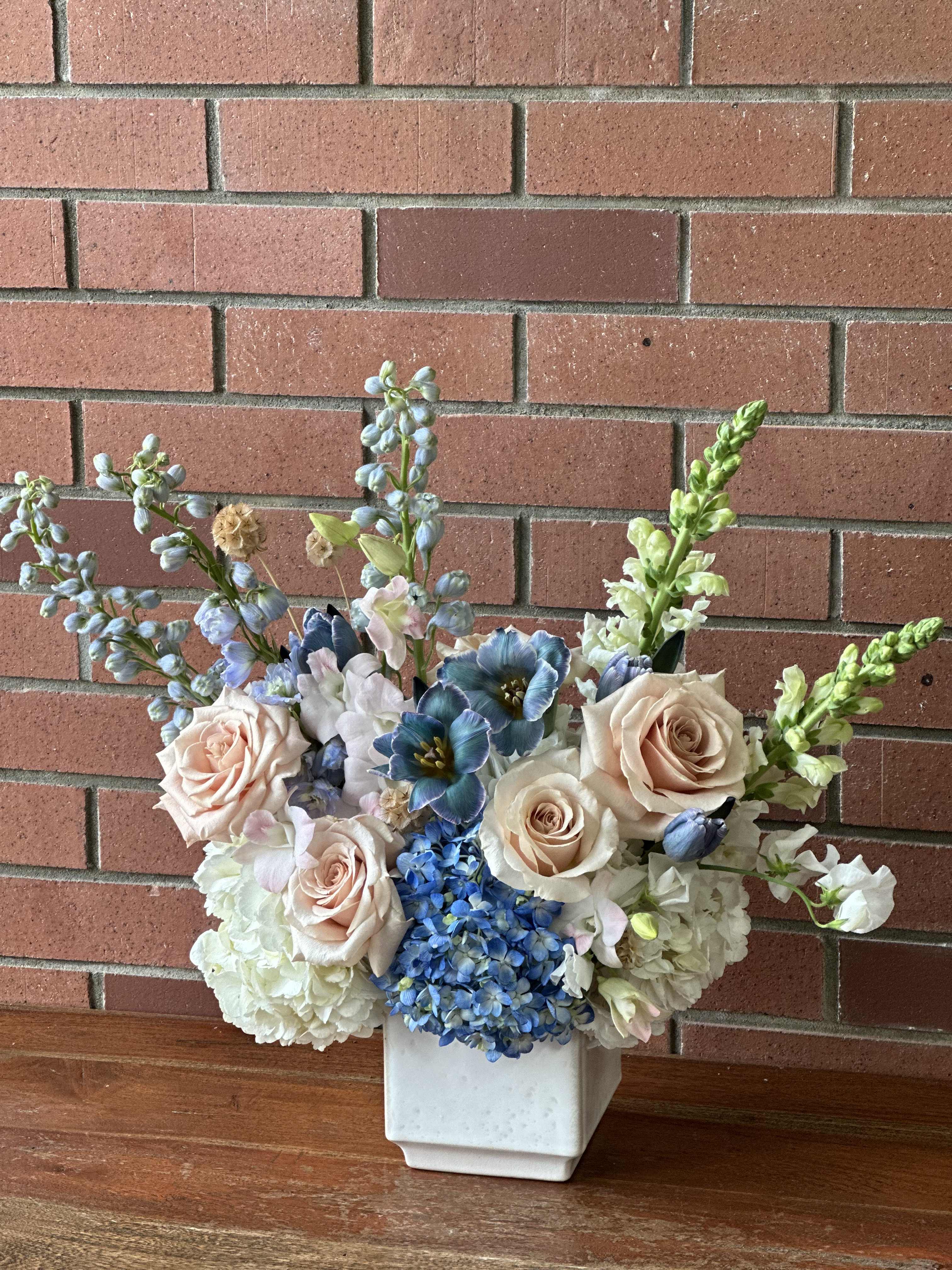Something Blue  - Send a fresh arrangement of blue and white hydrangeas, blue fresh tulips, blue delphinium, beige/blush roses, white snap dragons, white sweet pea, accented with blush dendrobium orchids designed in a modern ceramic vase 