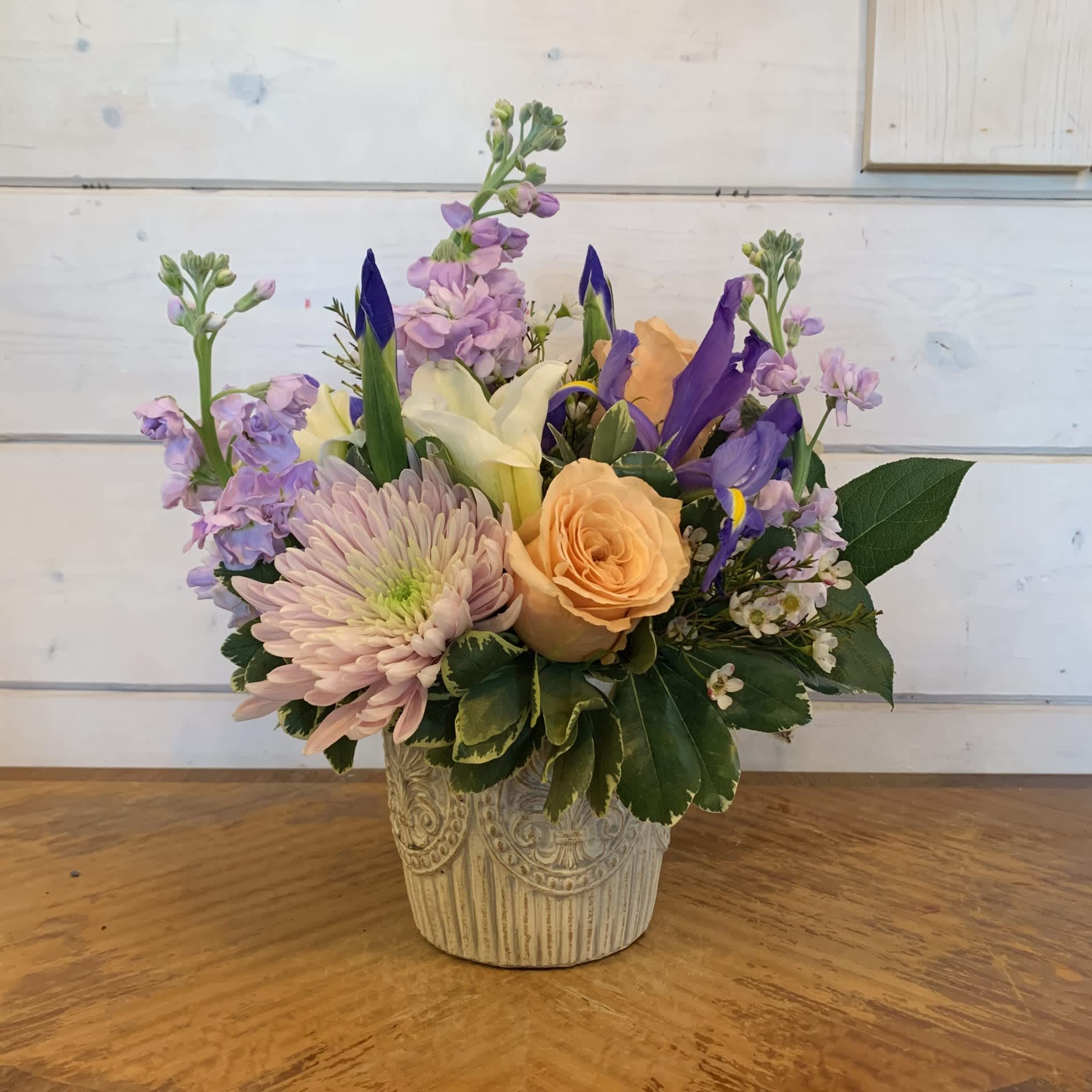 Elegant Spring Bouquet  - Pale apricot roses, lavender stock, white lily, linnet cremon chrysanthemums, and iris arranged in a small medallion pot with seasonal white wax. Standard version is approximately 10in tall and 10in wide. 