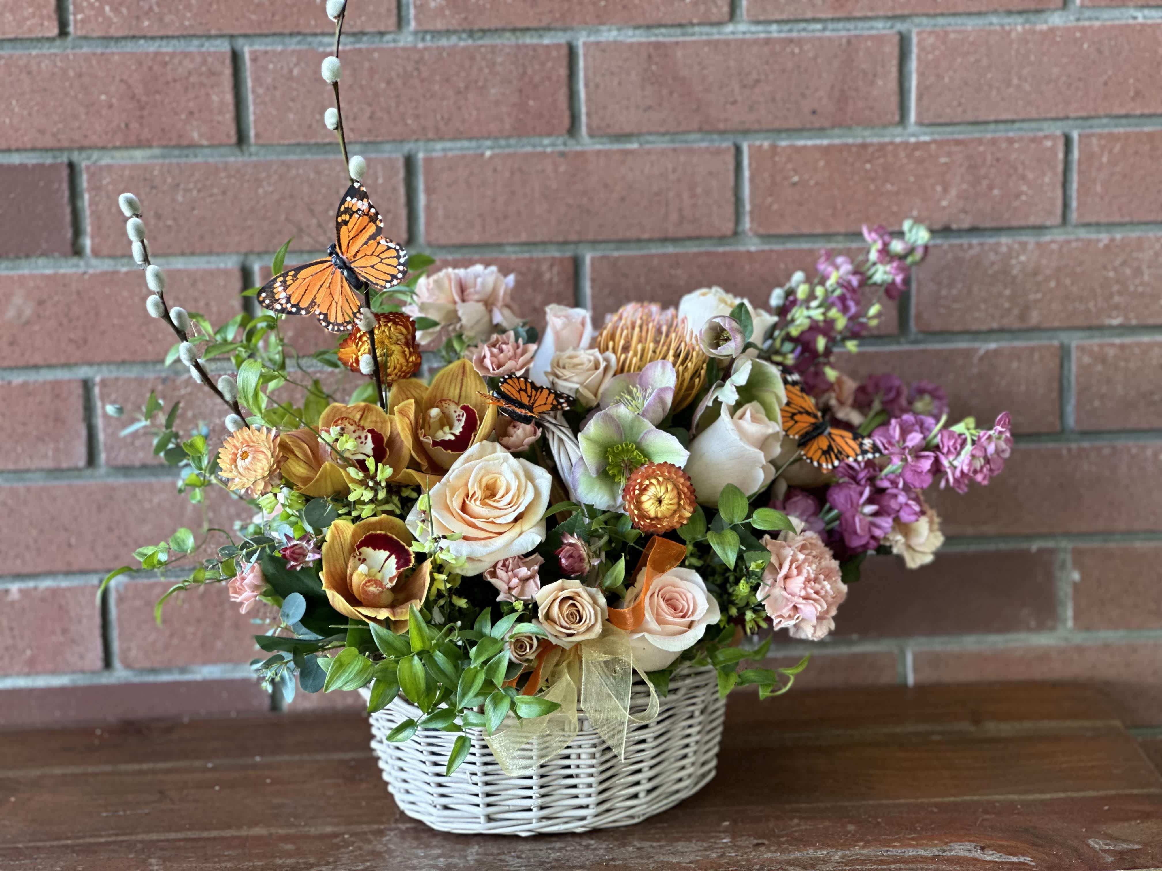 Fluttering Monarch Garden Basket  - Send a fluttering garden basket with Monarch butterflies - filled with orchids, roses, Hellebore, stock, carnations, pin cushion protea, strawflowers, spray roses and more. Accented with yellow and orange chiffon ribbons in a basket with a liner 