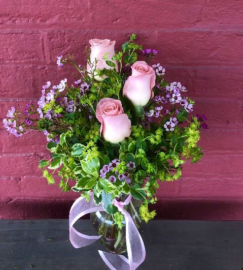 Looking Lovely 3 Pink Rose Vase - A sweet gift indeed. Three pink roses adorned with various greens and fillers. Topped off with a cute pink shoestring bow, tied around a clear glass vase.  Different colored rose available upon request.