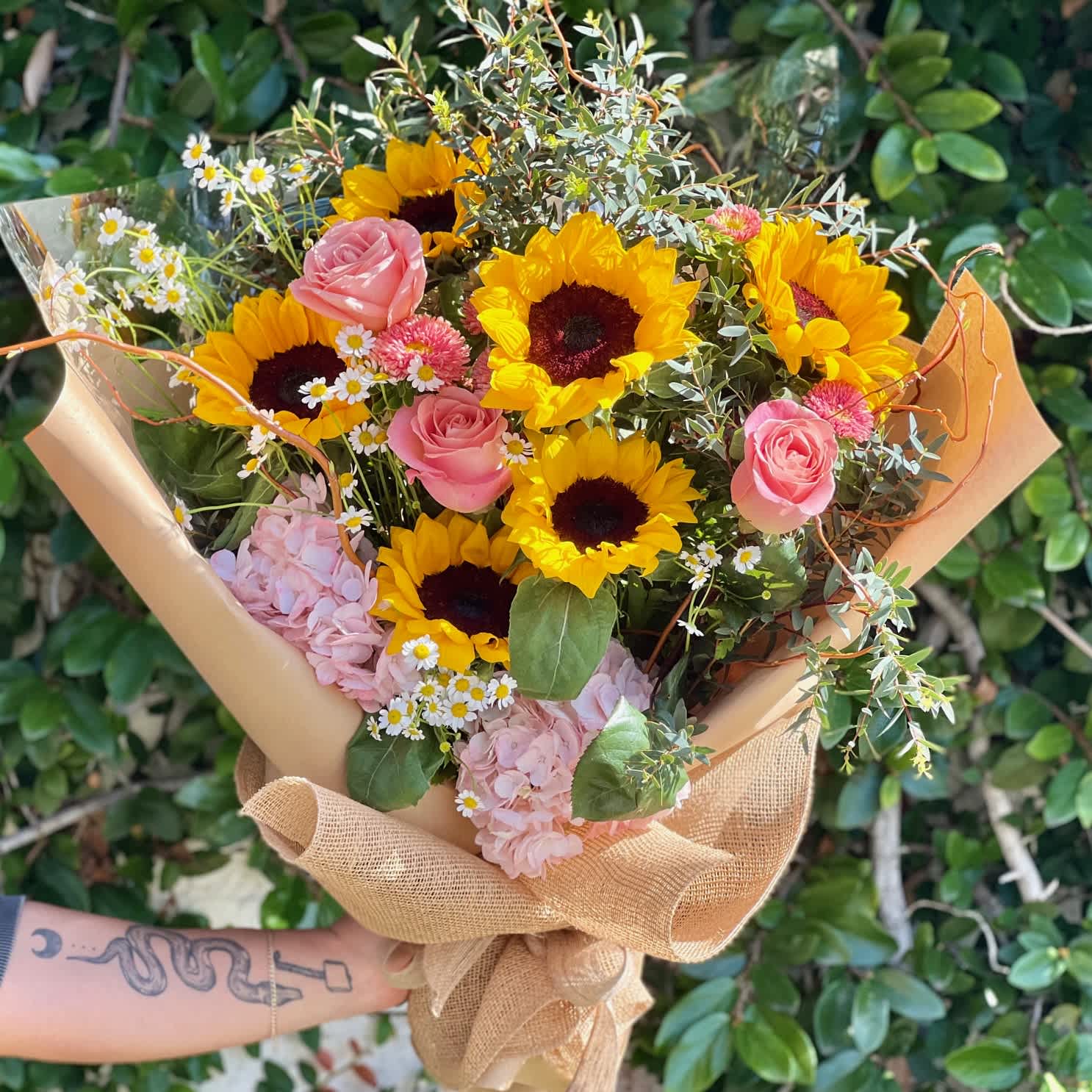 Sunflower Sunshine Bouquet - This radiant bouquet features a stunning mix of sunflowers, chamomile, pink roses, pink hydrangeas, peach Matsumoto flowers and more in a rustic burlap. Perfect for gifting a burst of sunshine!  NOTE: This bouquet DOES NOT include water tubes or a vase. It is wrapped with cellophane and brown paper and needs to be placed in water immediately after receiving.