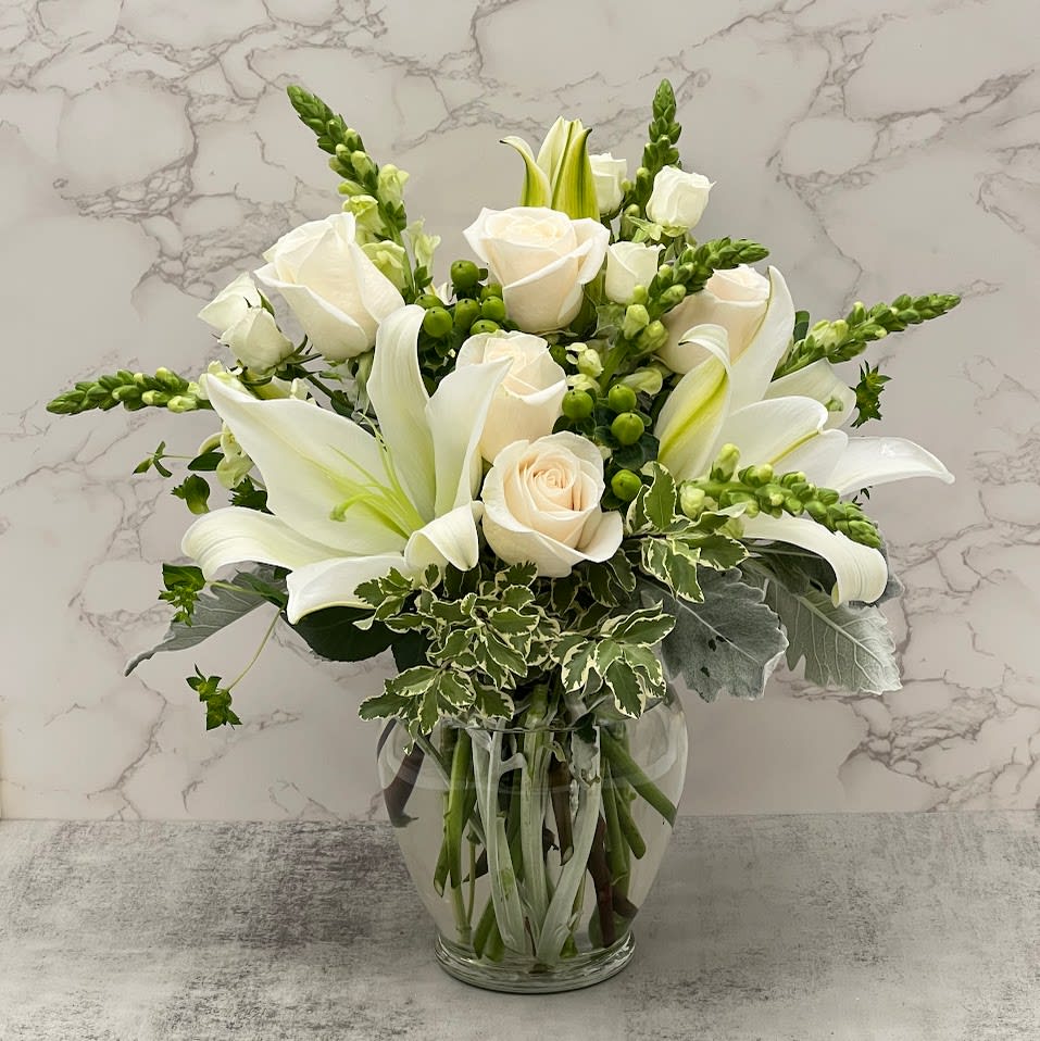Glamorous White  - All White elegant bouquet with lilies, white delicate roses, tall snap dragons, cute mini spray roses, and greenery to tie it all into a crystal-clear vase to make a statement of elegance and class. 