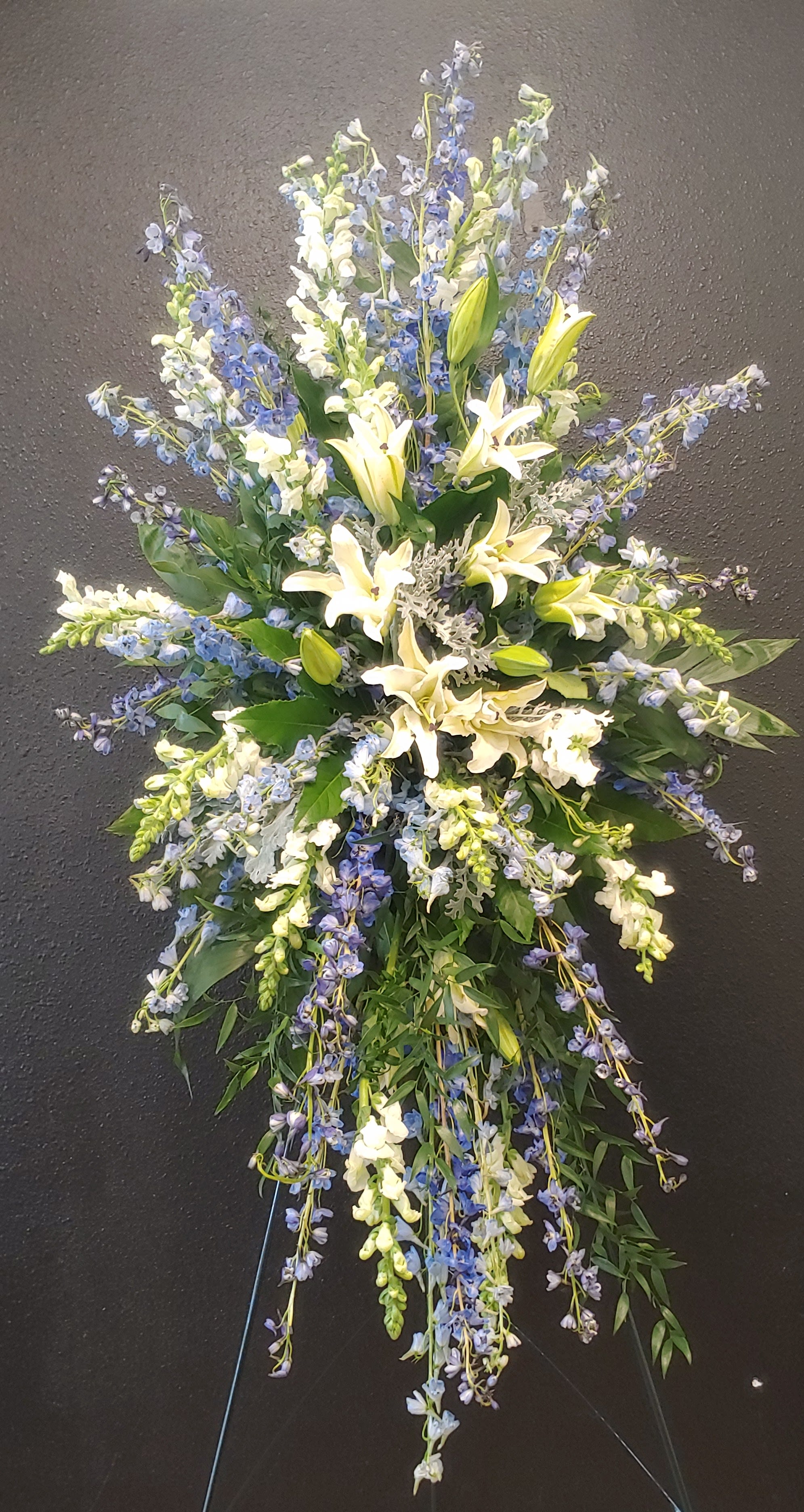 Heavenly Spray  - Sending this light Blue and White standing spray for a funeral expresses your most sincere sentiments and condolences to family and friends. Lovely blue delphinium and White Lillies, along with white snapdragons are accented with lovely greenery and situated on a tall easel stand.