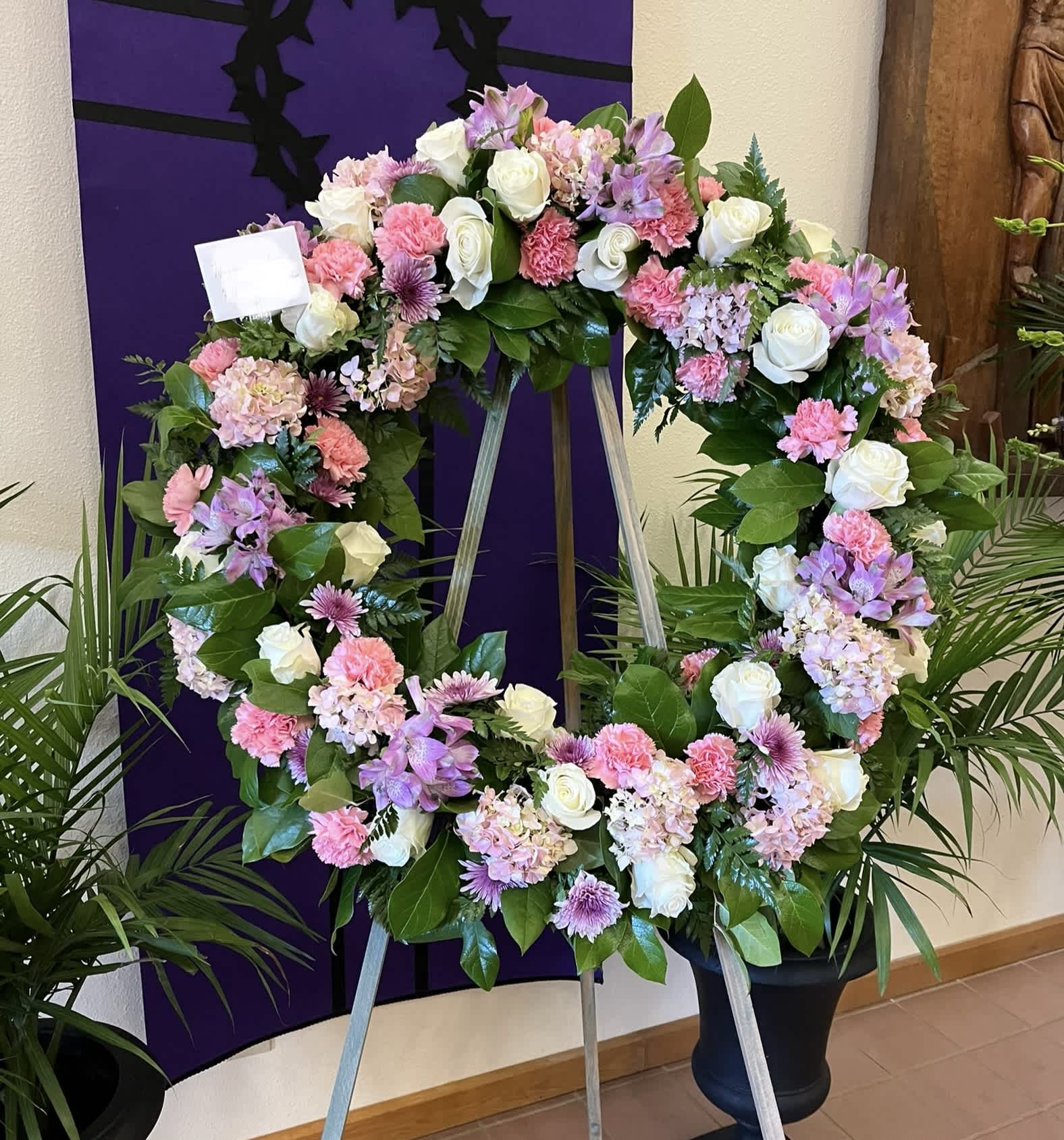 White Pink Lavender Wreath  - Sympathy wreath in a stand in a soft feminine color palette. Individual varieties may change according to season. Stands about 5 ft tall. Finished wreath is about 36” across.