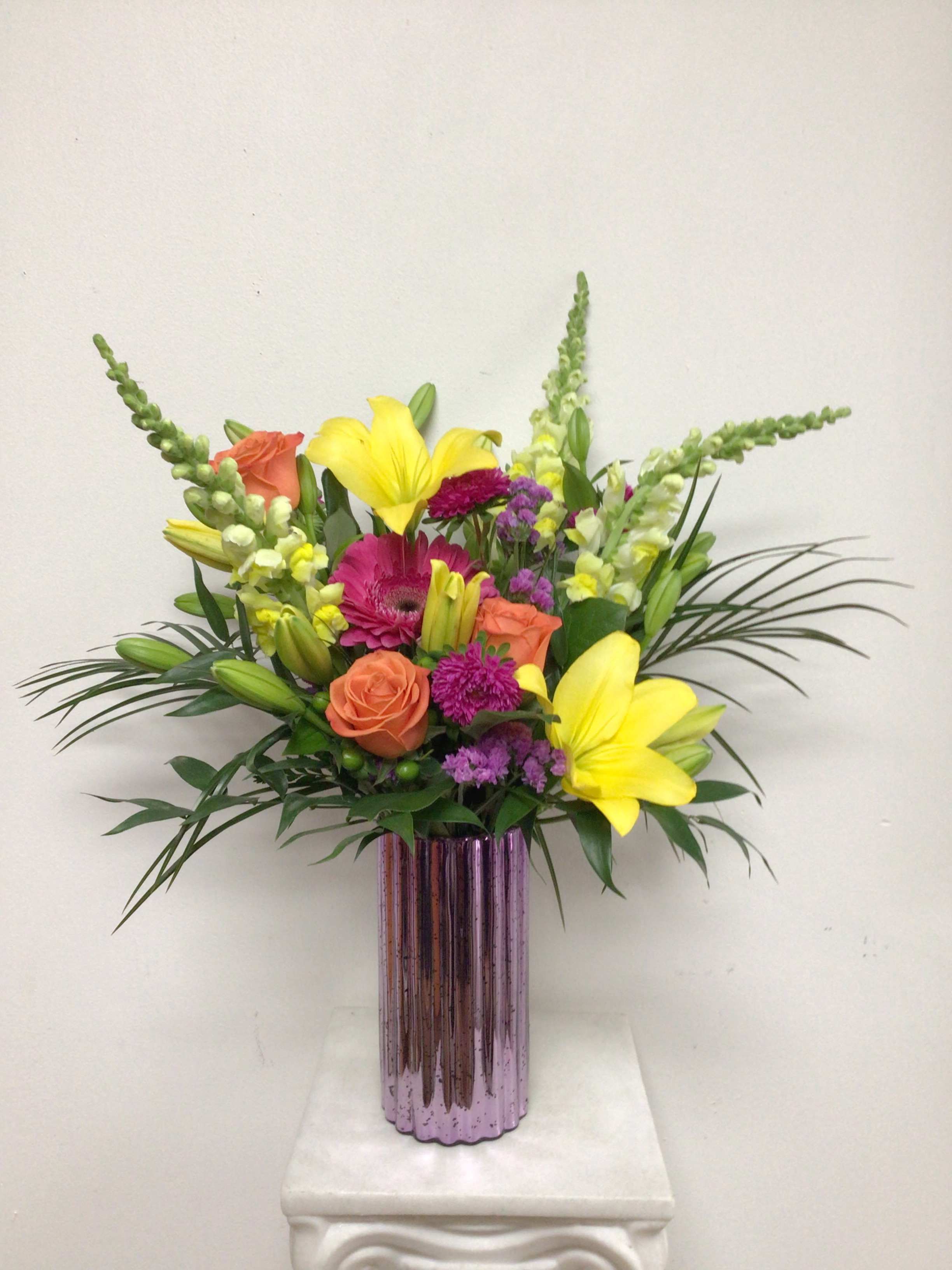 Color Blast - 8 inch lavender ribbed Mercury glass vase is arranged with yellow lilies, hot pink gerbera daisies, orange roses, statice and mixed greenery. Approximate size is 20 inches tall by 15 inches wide