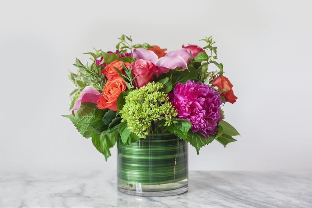 Celebration Collection - Market Size  - A glass, leaf lined cylinder with hot, hot colors for Summer. Vibrant Pink Peonies and Calla Lillies, Mini-green Hydrangeas, Orange and Pink Roses and a power packed combo of scented herbs with Rosemary and Mint; how can you resist? Our designers know the way to someone's heart and mind because they live and love flowers every day. And if you are ever on Market Street in Leesburg, stop by and meet our team.  Upgrade to Premium for 1 additional Peony, 3 additional roses, 3 additional mini green hydrangea and greenery and herbs. Arrangement will be approximately 16&quot; tall and 10&quot; wide. 