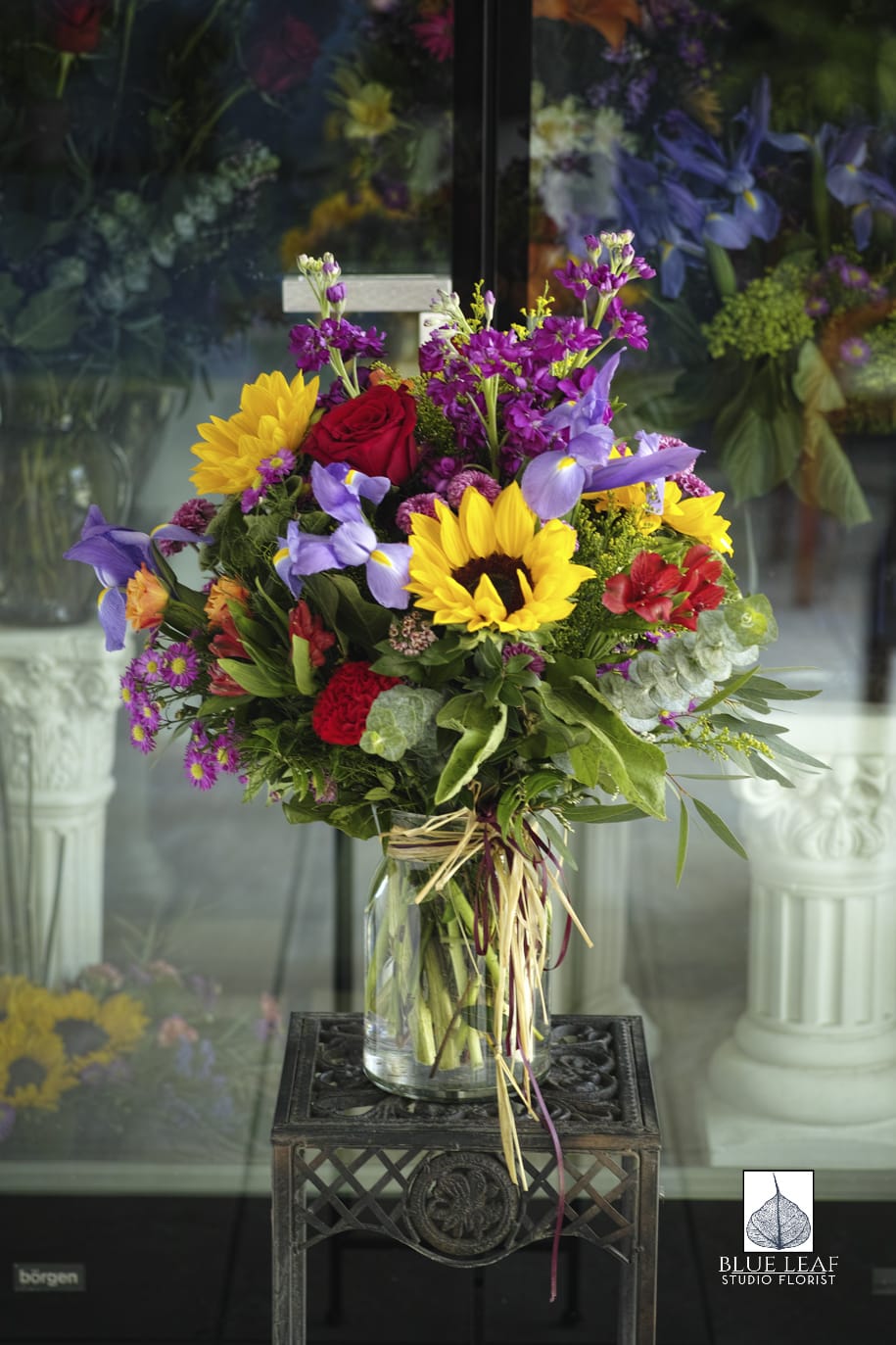 Summer Fun - A fun, uplifting bouquet of sunflowers, purple iris, red roses, stock, and other filler flowers arranged in a clear glass vase.