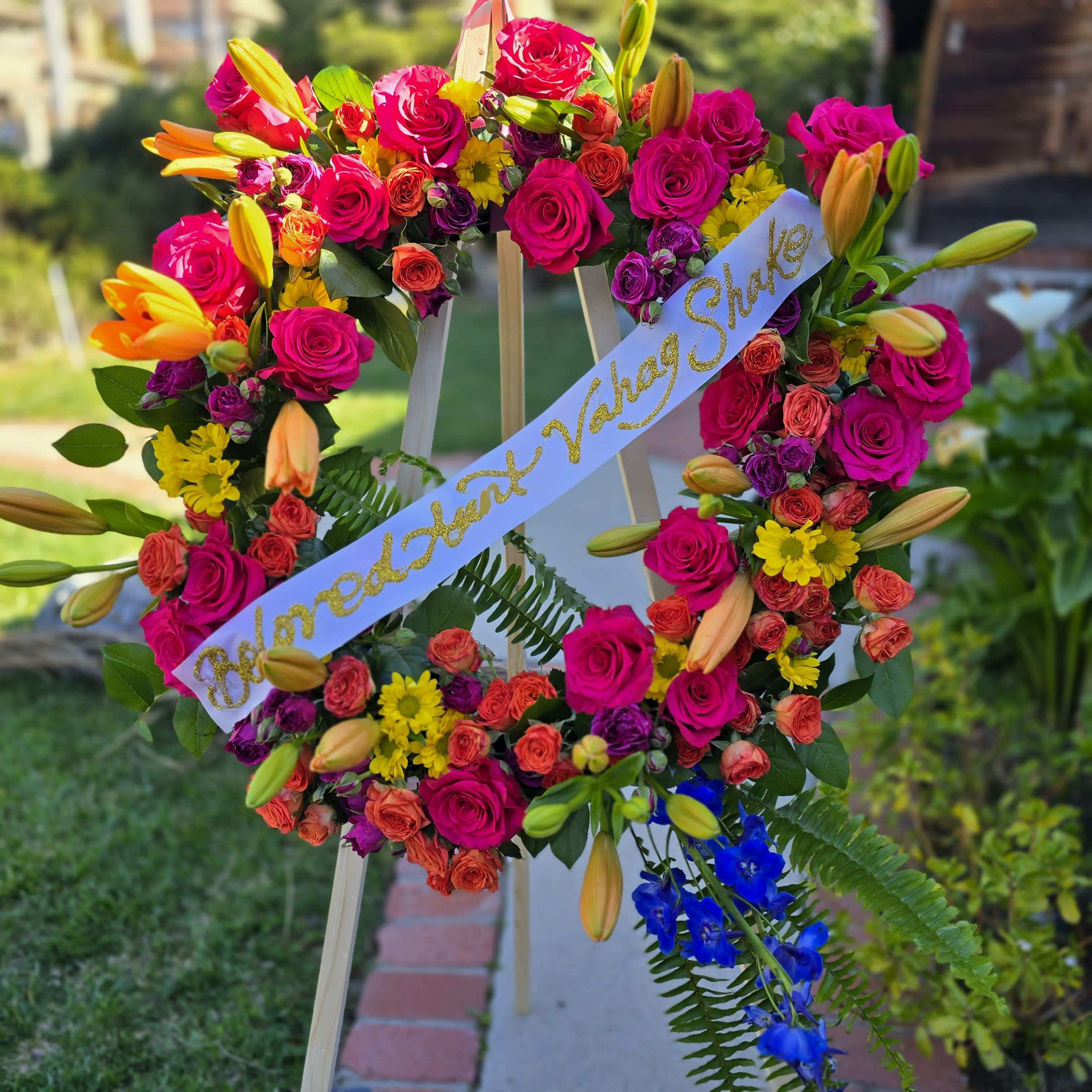 Colorful Serenity - a warm &amp; bright funeral spray, with orange lily, hot pink rose orange spray roses, purple stock, and yellow daisy. This wreath also has a blue delphinium and fern tail.