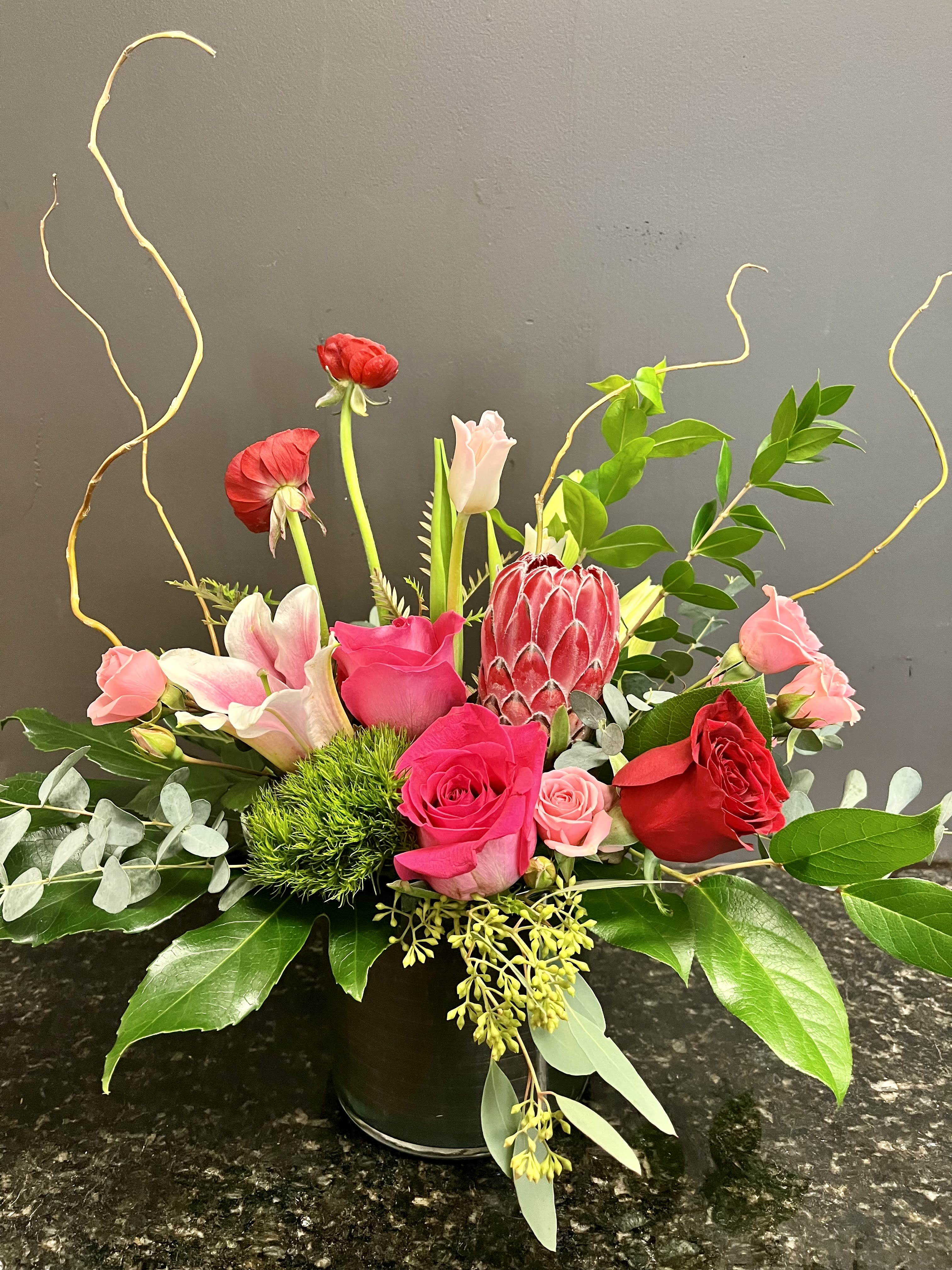 Aphrodite - a stylish mix of premium pink and red flowers including roses, lilies, tulips, ranunculus and accented with fancy greens in a glass cylinder. 
