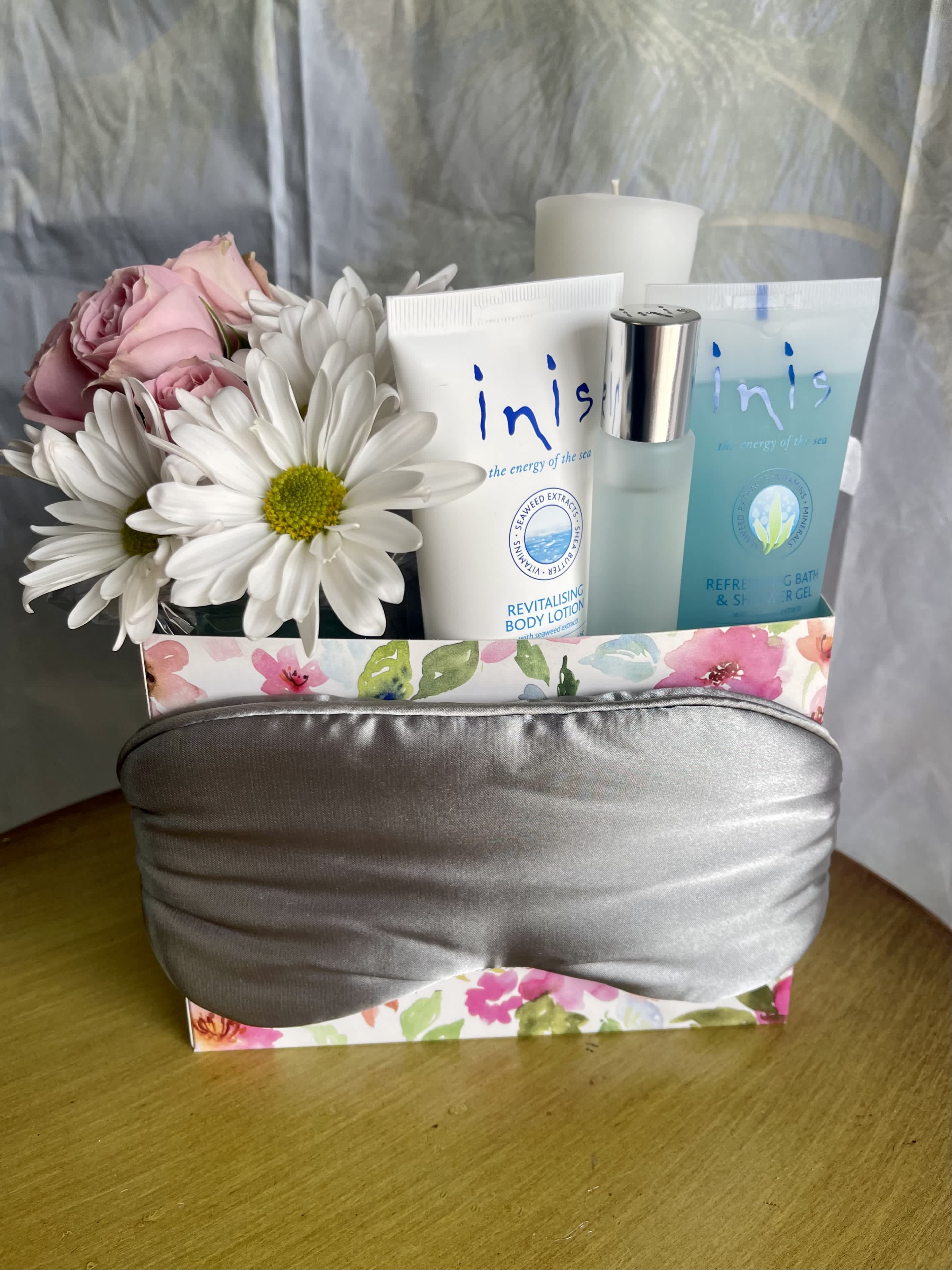 Peace and Pampering - A blooming box of relaxation for her! Gift set includes a small arrangement of fresh flowers, shower gel, lotion, and cologne by Inis, a votive candle by Tyler, a bath sponge, and a sleep mask! A thoughtful gift that promotes self care arrives in a floral printed box, wrapped in cellophane, and adorned with a ribbon that makes it a beautifully presented gift.