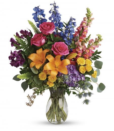 Colours Of The Rainbow - A colorful sympathy arrangement featuring a blend of vibrant blooms arranged into a tall clear glass vase with fresh mixed greenery.