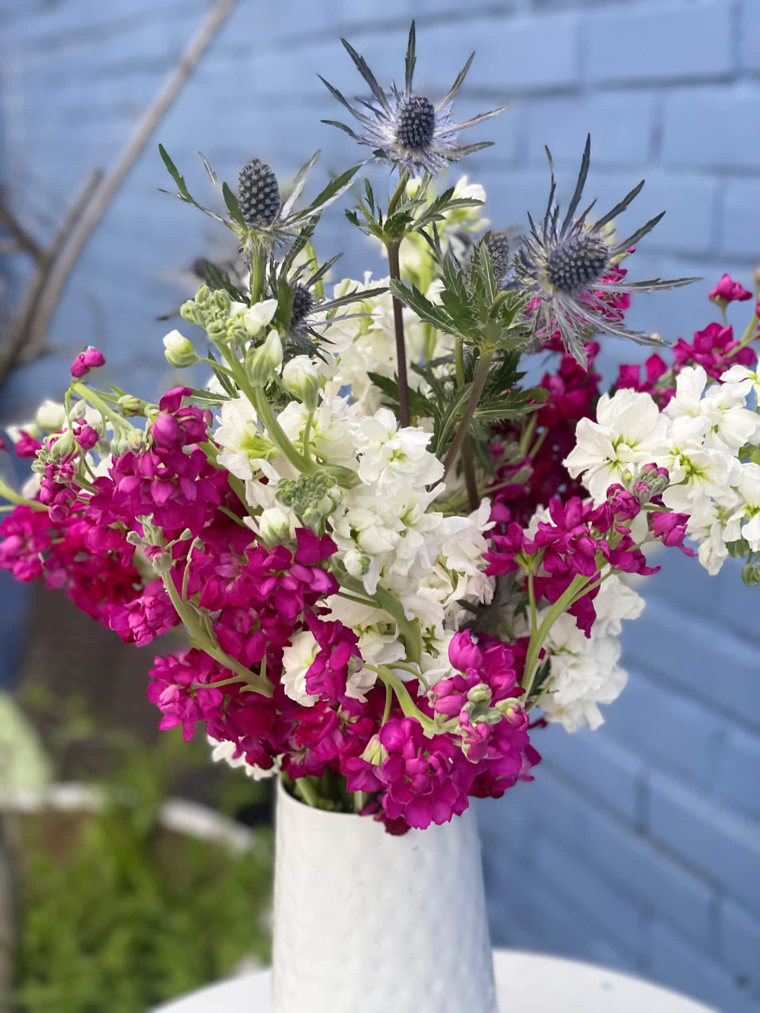 Raspberry Cream - This simple arrangement is packed with blooms and filled with fragrance! The white and raspberry stock spilling out, mixed with the navy blue of the Sea Holly, make this piece a stunning addition to any home and is sure to smell fresh for days!