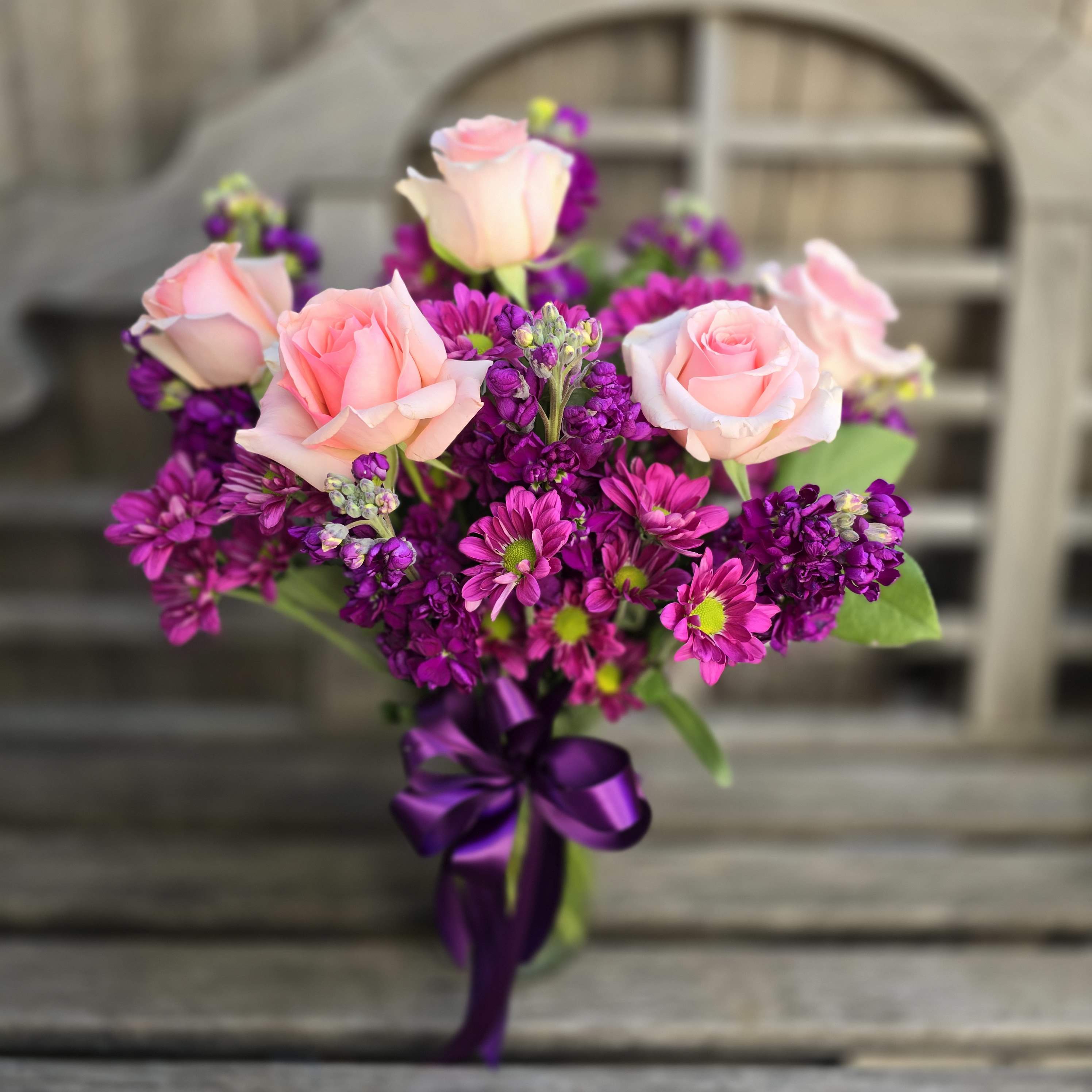 Sweet As Can Be - Sweet as can is a lovable bouquet featuring pink roses, purple daisy, and purple stock presented in a clear glass vase!  This arrangement suits many occasions and can easily be upgraded to the perfect birthday gift with a birthday theme Balloon, and Birthday Theme Enclosure card.  Our standard size is designed around 5 focal roses Our Deluxe is Designed around 7 focal roses Our Premium is designed around 10 focal roses