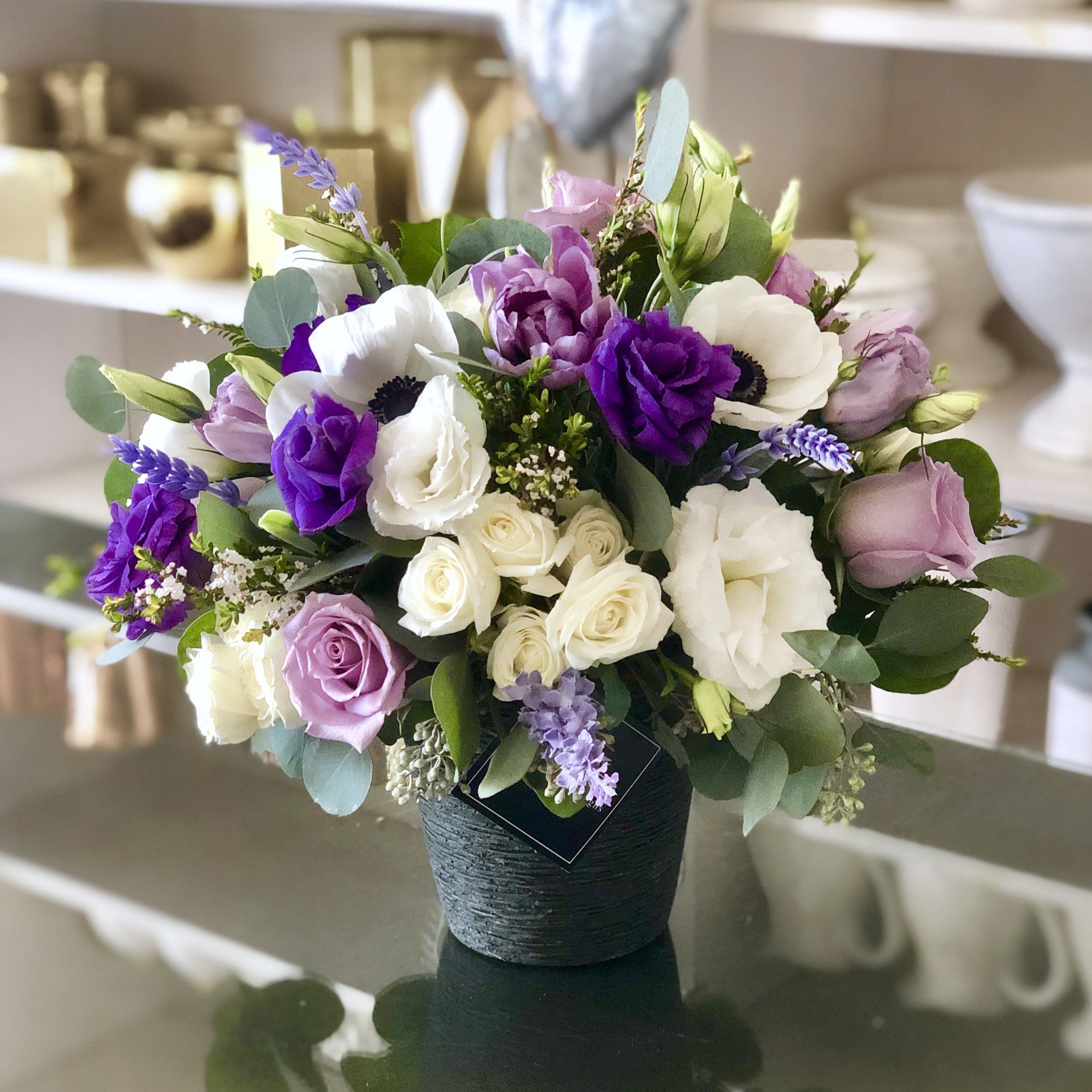 Sassy - A lively, bold, and full of spirit arrangement is best described our Sassy Bouquet. A textured clay pot filled with Purple and White Lisianthus, White Spray Roses, White Roses, Purple Stock, Lavender Roses, Seeded Eucalyptus, White Anemones.   (Please note when Anemones are not in season we will use other premium florals as a substitution)