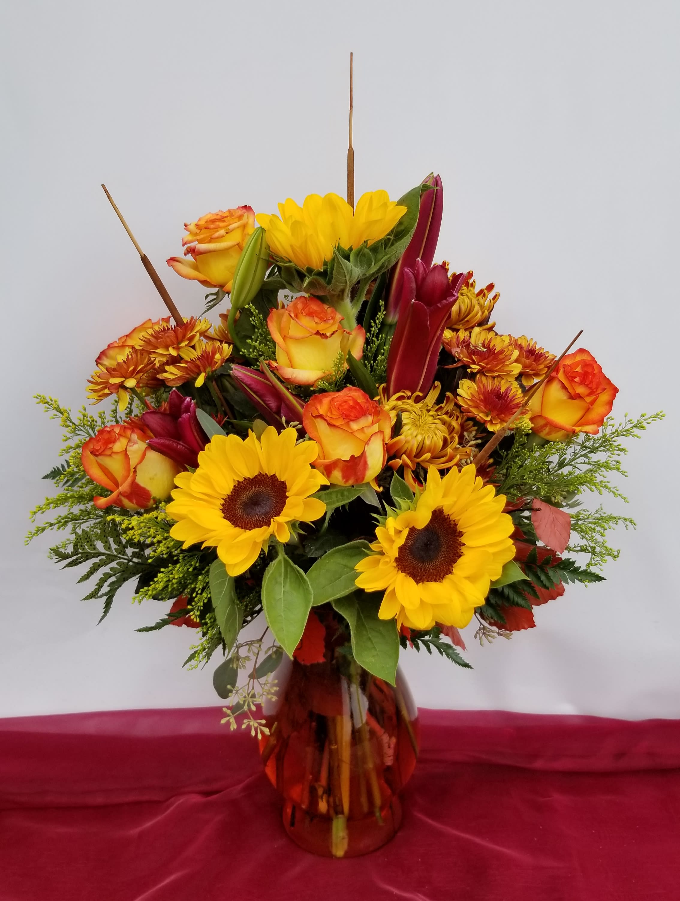 Autumn Radiance Bouquet - This warm array of yellows, oranges and reds is fitting for and Fall Festivities! With its' lovely mixture of roses, lilies, daisy mums, and sun flowers, there is no doubt that is will bring joy to someones day. It is accented with Solidago, Eucalyptus and Fall accessories (dried autumn leaves, sticks, etc.) This is arranged in a clear glass vase (not red). The photo shown is the Deluxe version. 
