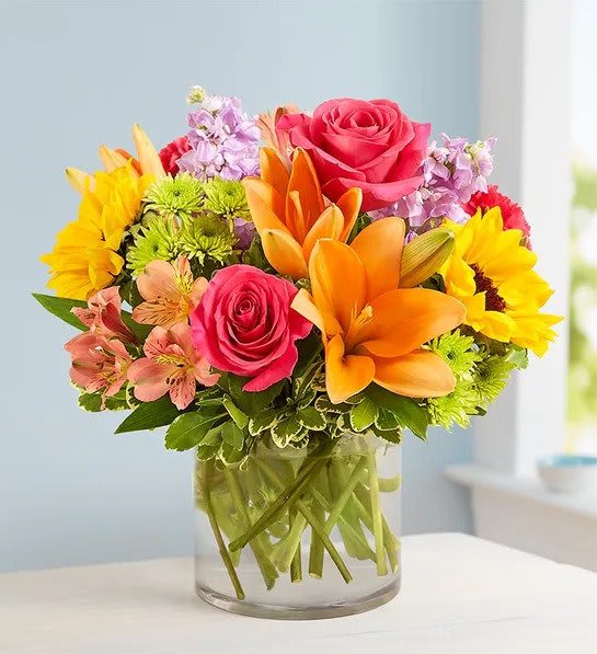 Floral Embrace - Like a warm embrace, our vibrant flower bouquet delivers your sentiments to someone special. A rich gathering of yellow and orange blooms, with pops of bright pink and purple, it's more than a gift…it's a way to express how you feel inside.  - All-around arrangement with hot pink roses and carnations; orange Asiatic lilies and Peruvian lilies (alstroemeria); yellow sunflowers; lavender stock; purple monte casino; green Athos poms; accented with assorted greenery - Premium arrangement measures approximately 16&quot;H x 14&quot;W - Standard arrangement does not include monte casino; measures approximately 14&quot;H x 13&quot;W - Artistically designed in a clear glass cylinder vase; large vase measures 6&quot;H; medium/small vase measures 5&quot;H - Our florists hand-design each arrangement, so colors, varieties and container may vary due to local availability - To ensure lasting beauty, lilies may arrive in bud form, and will fully bloom over the next few days - Sunflowers may arrive with light or dark centers