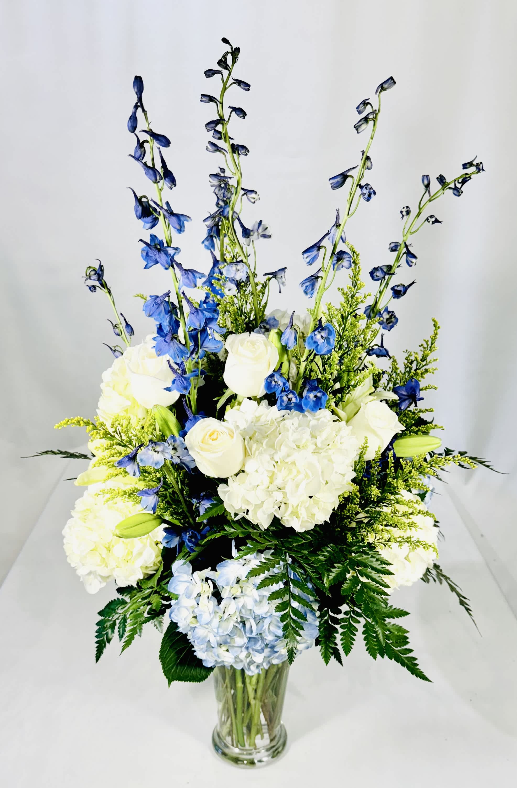 Blue & White Hydrangea Rose Arrangement Blue & White Hydrangea Rose Arrangement - An elegant vase of blue and white hydrangeas, white roses accented with a pop of yellow solidago, blue delphinium and white lilies. Stunning arrangement with class and elegance for any occasion. 34" Tall