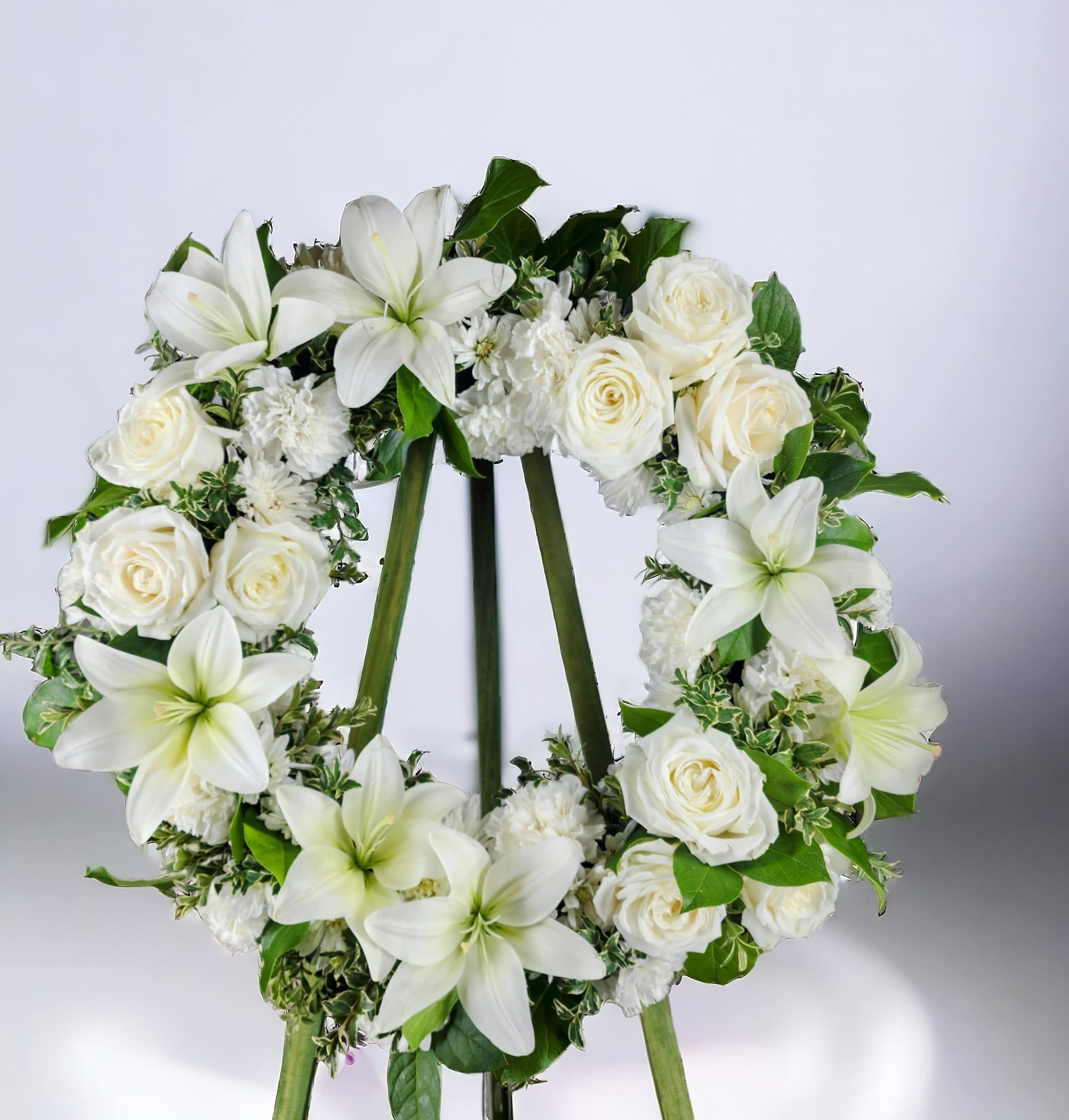 Elegant White Sympathy Wreath - This 18&quot; wreath is created using white roses, lilies, carnations and daisies on a bed of greens. A wire tripod stand is included.