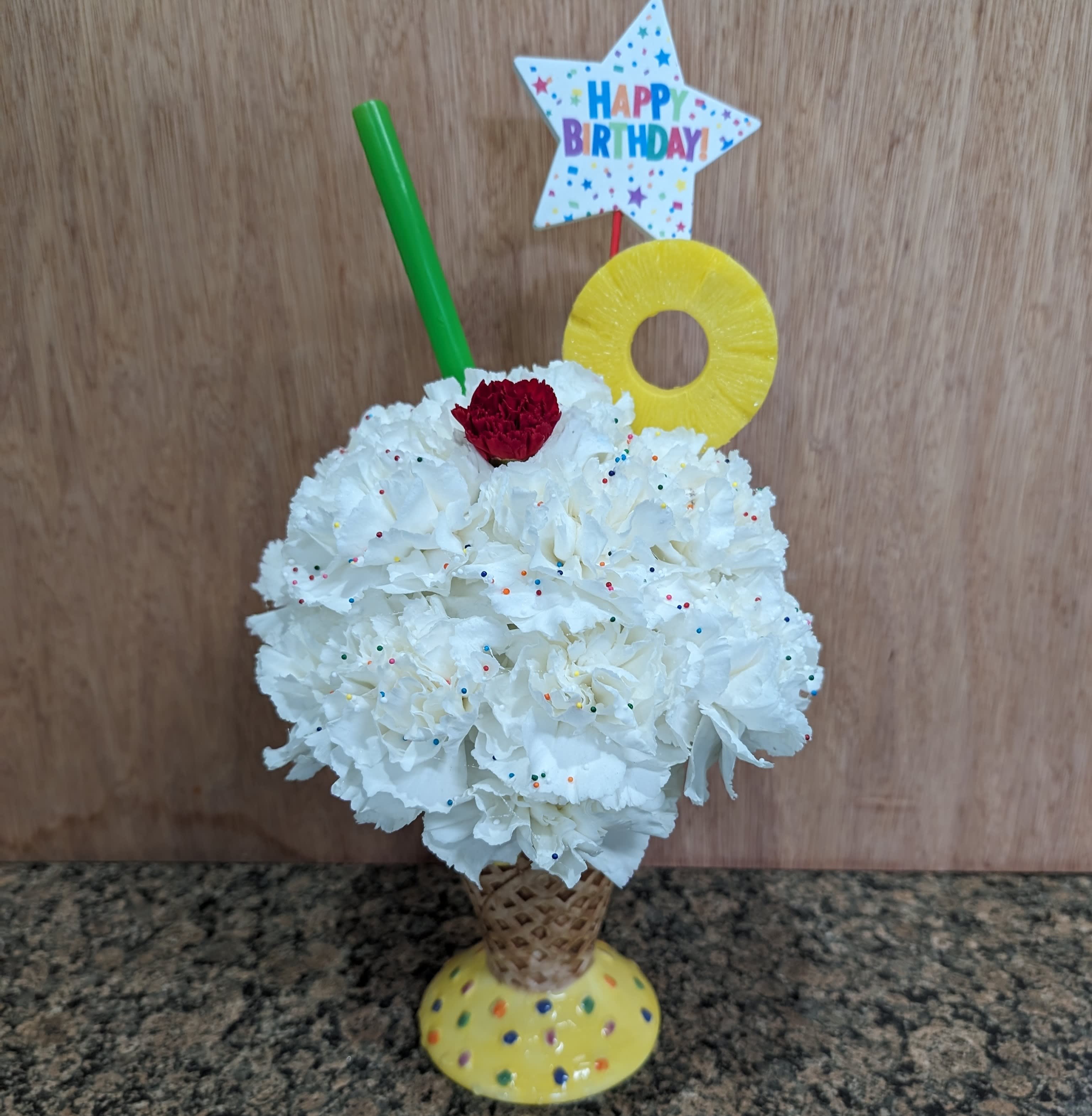 Flower cone sunday Arrangement - How festive is this?!? White carnations make up the ice cream arranged in a ceramic ice cream waffle cone. Adorned with a faux pineapple slice, sprinkles (or jimmies) straw and birthday card holder pick. This is sure to start the festivities off right.
