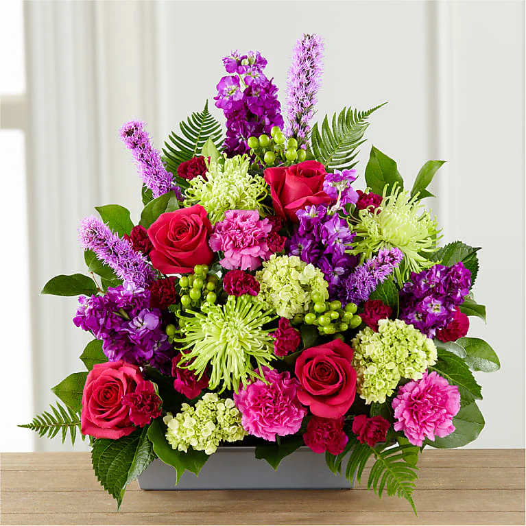 Warm Embrace Arrangement - A lovely way to say 'farewell,' this bright and beautiful arrangement expresses your sympathy through color, texture, and floral artistry. It offers comfort to grieving friends and family with hot pink roses, pink and fuchsia carnations, purple stock, green spider chrysanthemums, mini-hydrangea, and hypericum berries, all accented with sword fern and salal in a low rectangle container.