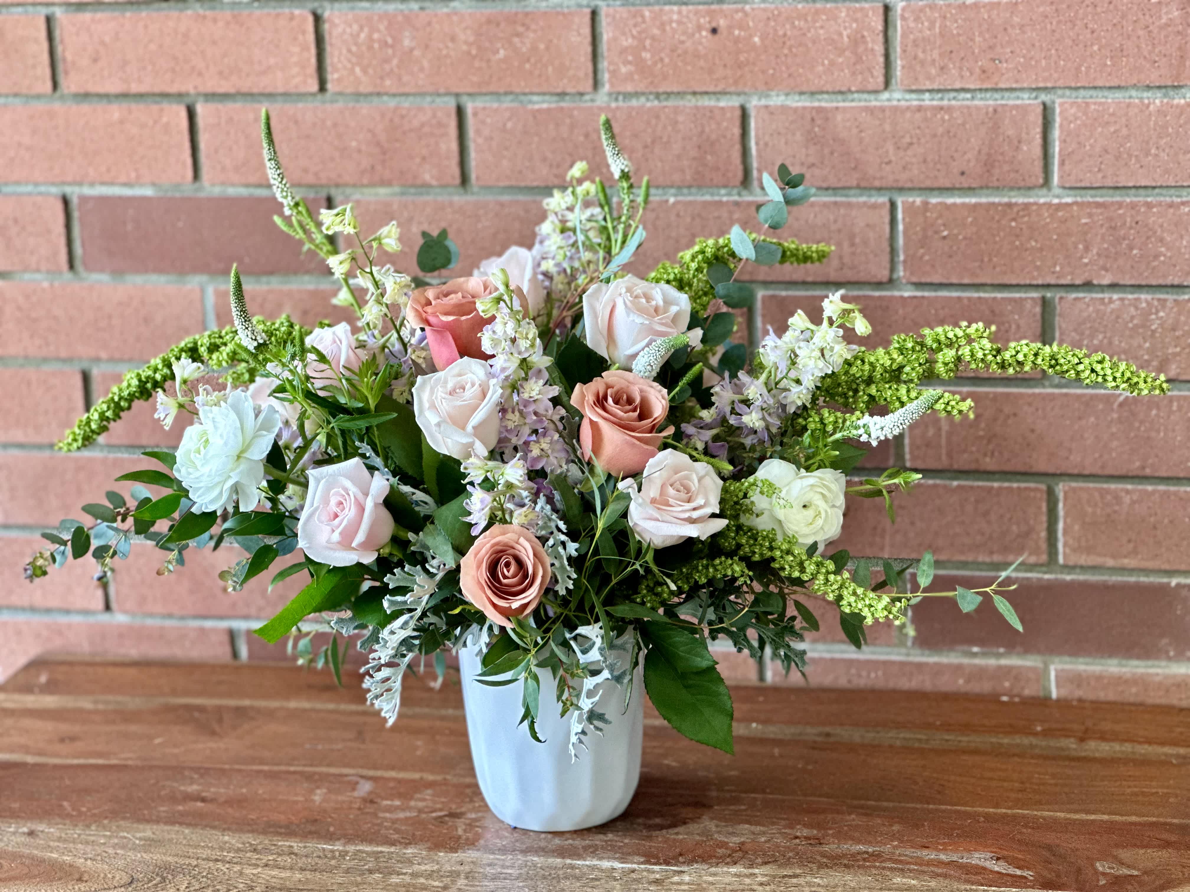 Whispering Petals Rose Bouquet  - A soft bouquet with blush and mauve roses, arranged in a white ceramic vase, is an elegant and delicate arrangement. The blush roses add a touch of romance and tenderness, while the mauve roses bring a subtle hint of sophistication. The combination of these shades creates a harmonious and soothing color palette.  To enhance the softness of the bouquet, wispy and lush greenery is incorporated. The greenery adds a natural and organic feel, creating a beautiful contrast against the delicate roses. The wispy foliage gently cascades, adding a touch of movement and grace to the arrangement.  Placed in a white ceramic vase, the bouquet exudes a sense of purity and simplicity. The clean lines of the vase complement the softness of the flowers, allowing them to take center stage. The overall effect is a stunning and serene bouquet that embodies elegance and tranquility.