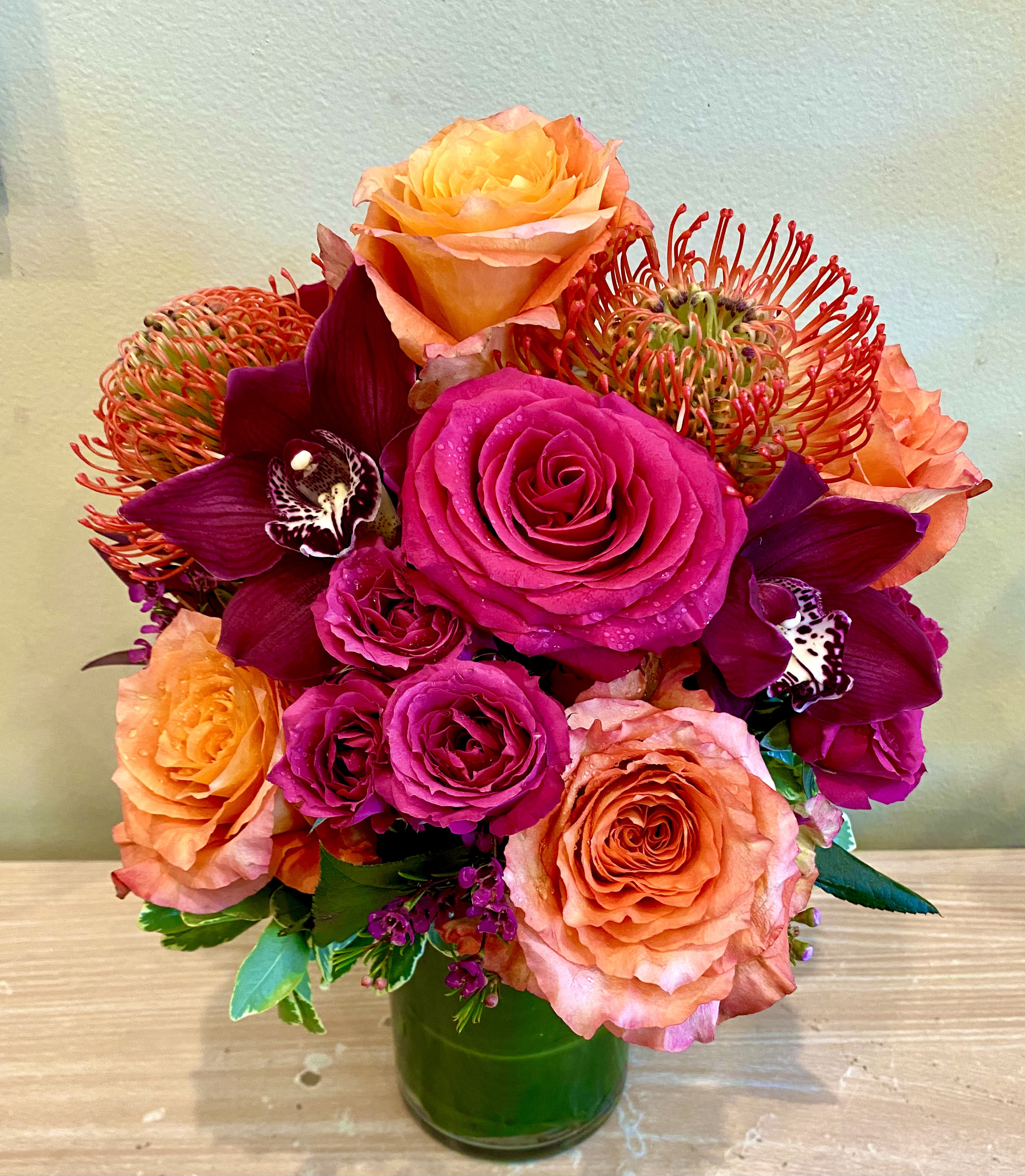 Warm Embrace - Deep hues of hot pinks, burgundy, coral &amp; orange roses, cymbidiums, and pin cushion protea add warmth and fire to this combination.