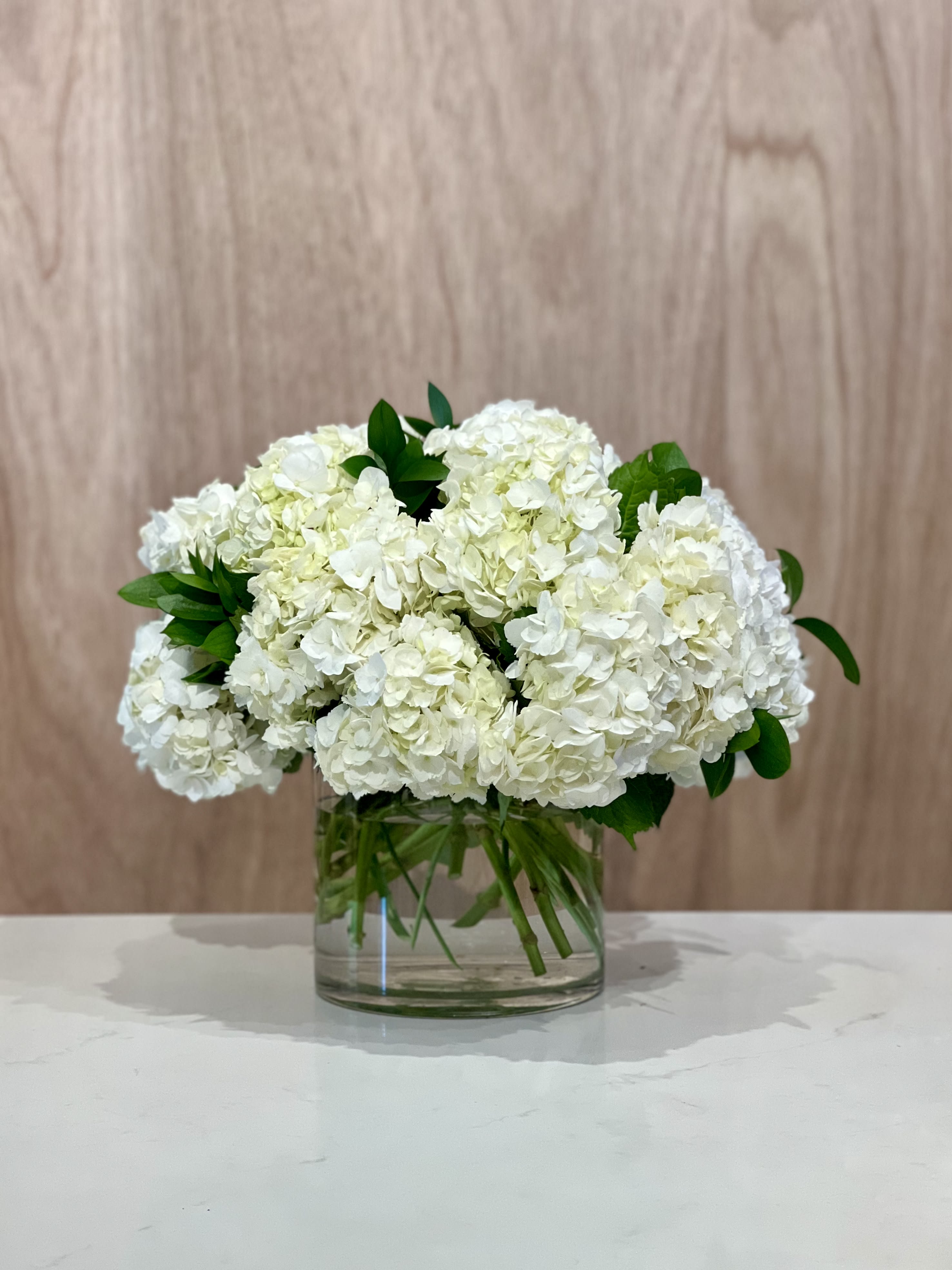 HYDRANGEA ARRANGEMENT - Embodying timeless sophistication, this exquisite design features a cluster of hydrangeas, carefully curated in varying sizes and hues. Nestled within an elegant glass vase, the arrangement exudes a refined and classic aesthetic.