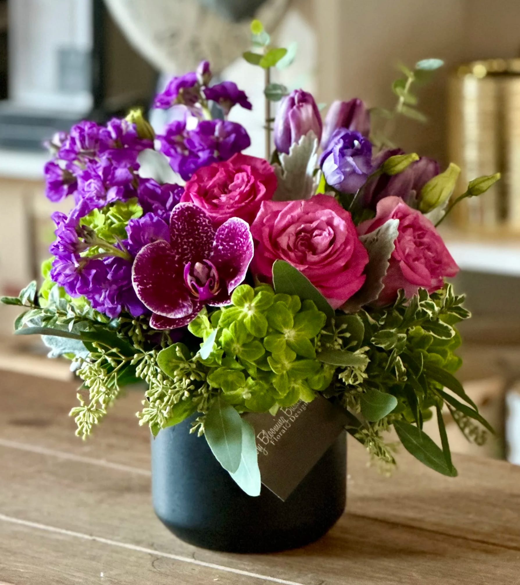 Xiomara - Introducing Xiomara, the stunning floral arrangement that is poised to captivate your family and friends with its unparalleled beauty. Immerse yourself in a world of jewel-tone colors and vibrant green hydrangeas, gracefully arranged in a sleek and sophisticated matte black vase. 