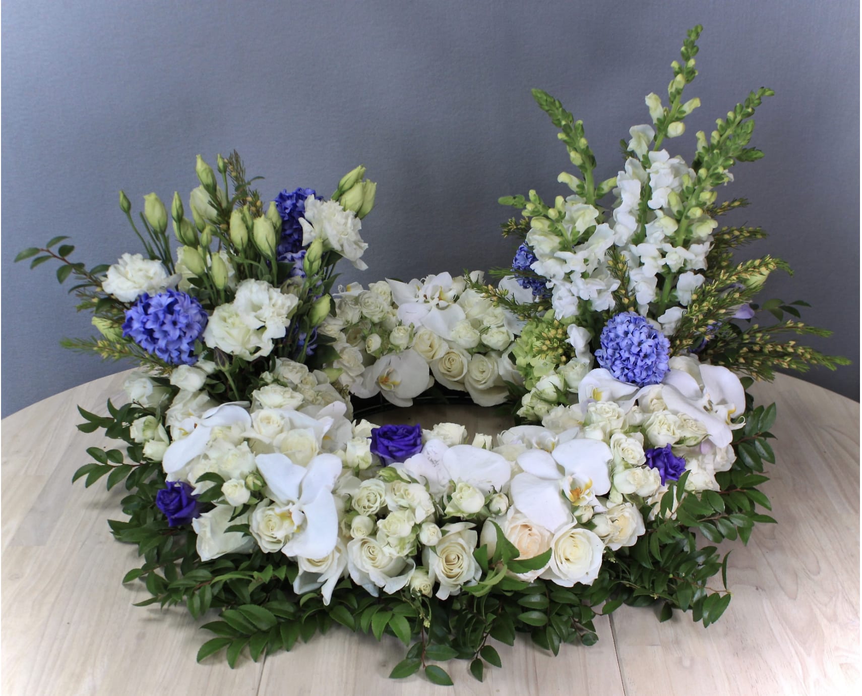 Urn Wreath in Blues  - This urn tribute wreath includes a mix of flowers, roses, and seasonal greens. In the special instruction let us know what colors you would like. Standard size is 30'', deluxe is 36'' and premium is 42''. Interior diameter is about 10'' for standard size.