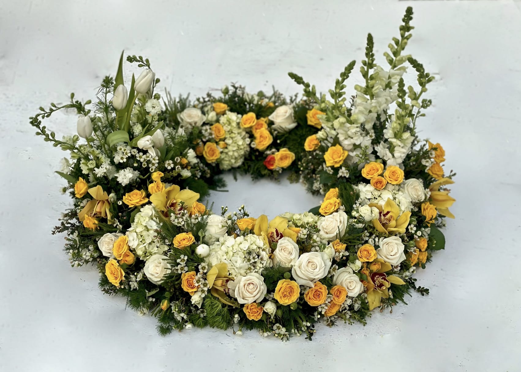 Urn Wreath in Yellows  - This urn tribute wreath includes a mix of flowers, roses, and seasonal greens. In the special instruction let us know what colors you would like. Standard size is 30'', deluxe is 36'' and premium is 42''. Interior diameter is about 10'' for standard size.