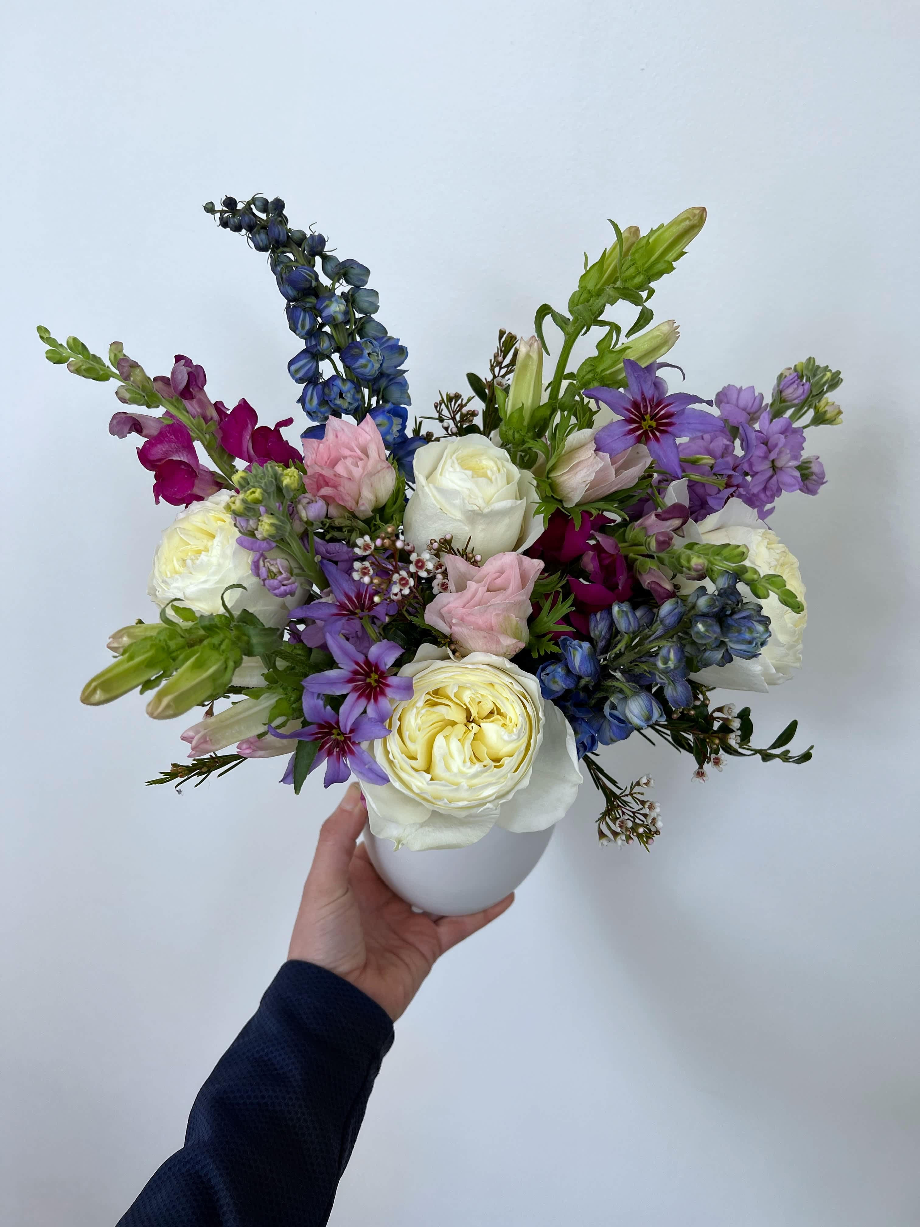 The Kimberly - Meet “the Kimberly”, a feminine variety of cream, pink, purple  and splash of blue florals. Arranged neatly in a white ceramic pot.