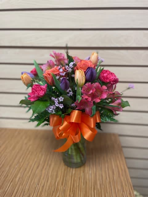 Burst of Citrus - Mixed cut arrangement with purple tulips, peach tulips, orange roses, hot pink alstroemeria, and hot pink carnations in vase with bow