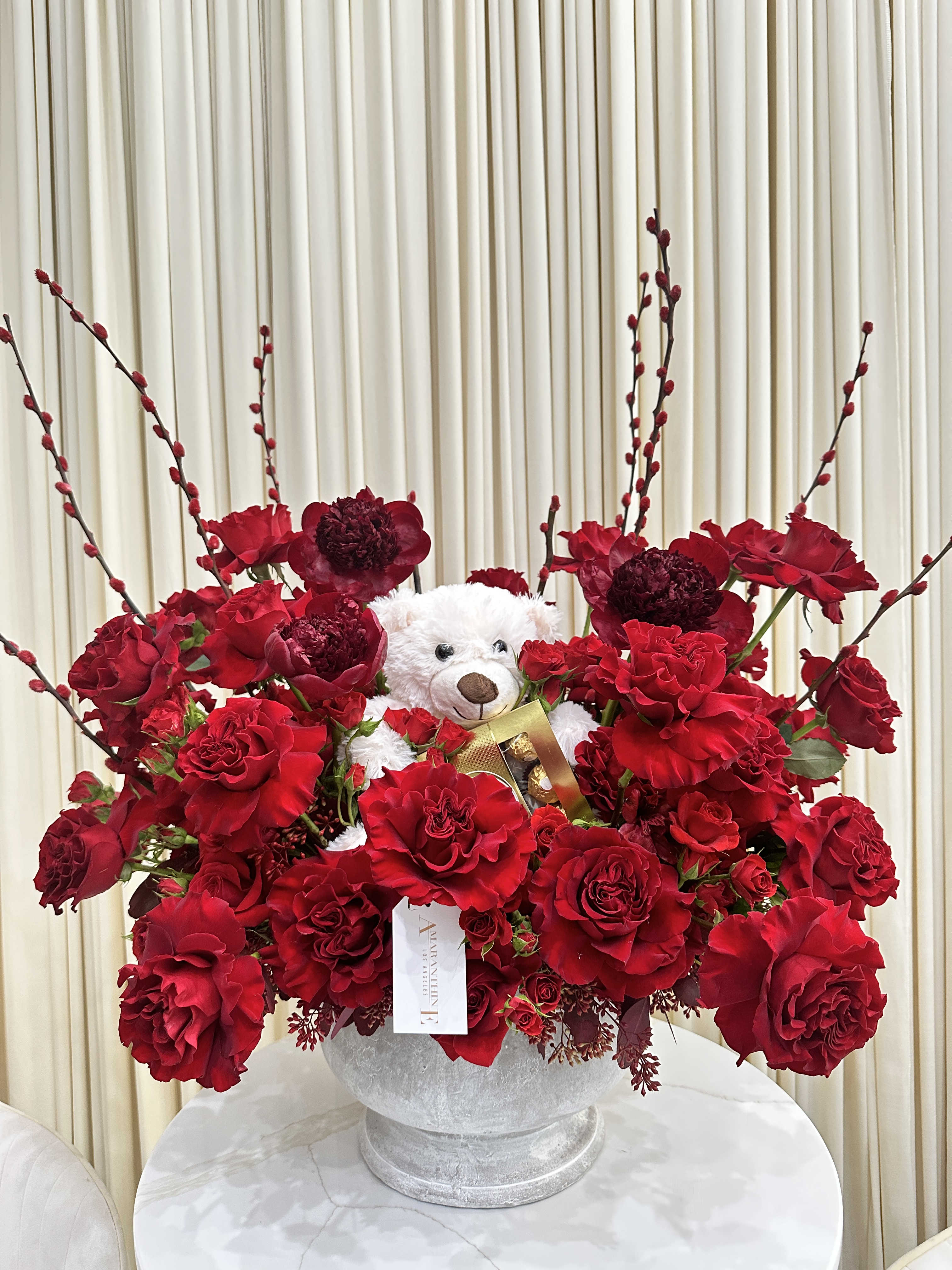 Crimson Cuddle - This delightful arrangement features a cuddly white teddy bear nestled amidst a vibrant bouquet of red roses. The plush bear, with its button eyes and stitched smile, brings a touch of whimsy to the classic elegance of the red roses. The roses add a pop of color and fragrance to any room.  This arrangement would be perfect for a variety of occasions, such as Valentine's Day, Mother's Day, or a birthday. It would also make a thoughtful and unique gift for any teddy bear lover.