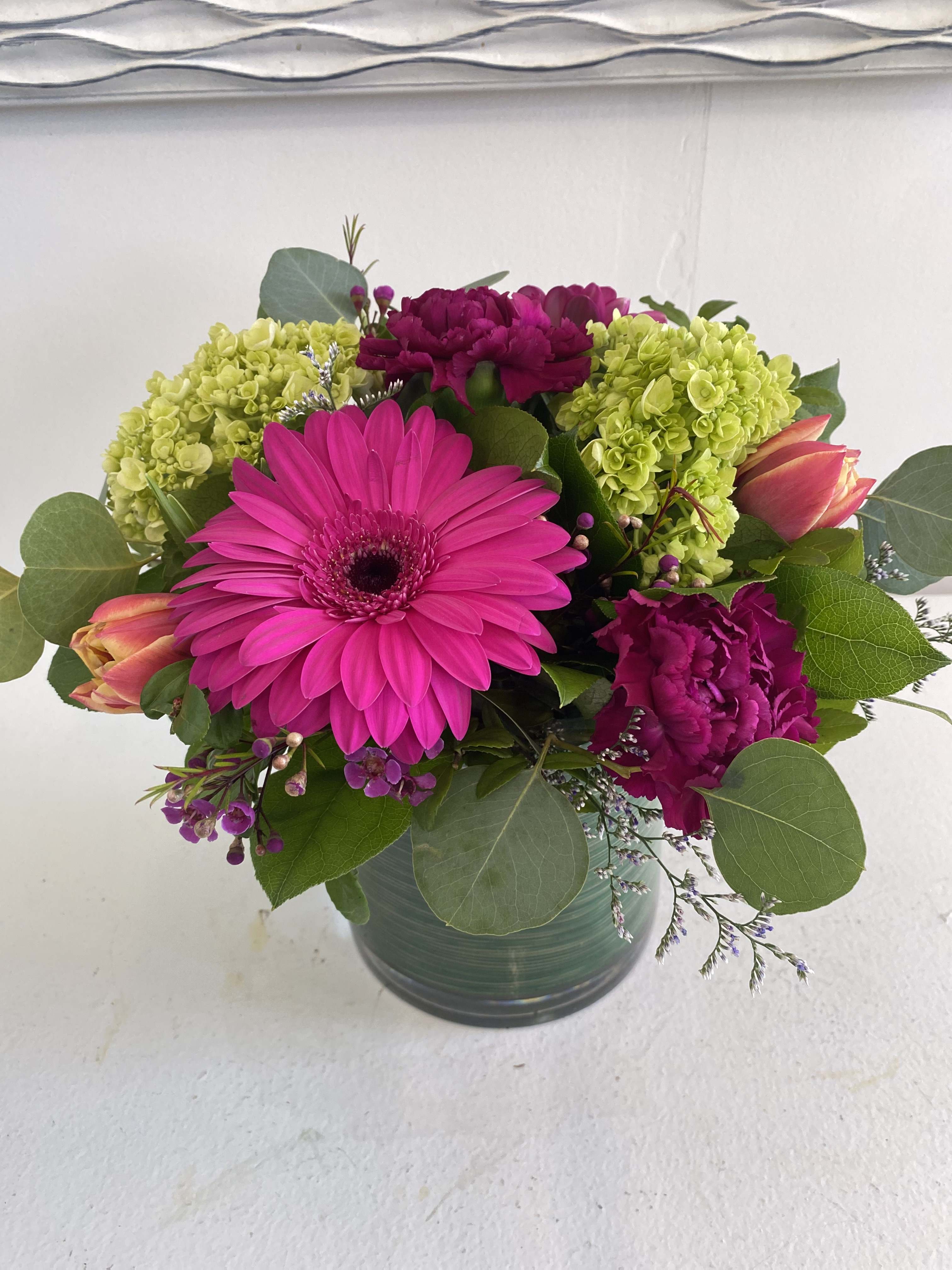 Mini Spring Blush - An eye-catching blend of fuschia, green, and purple blooms arranged into a glass vase with fresh mixed greenery.