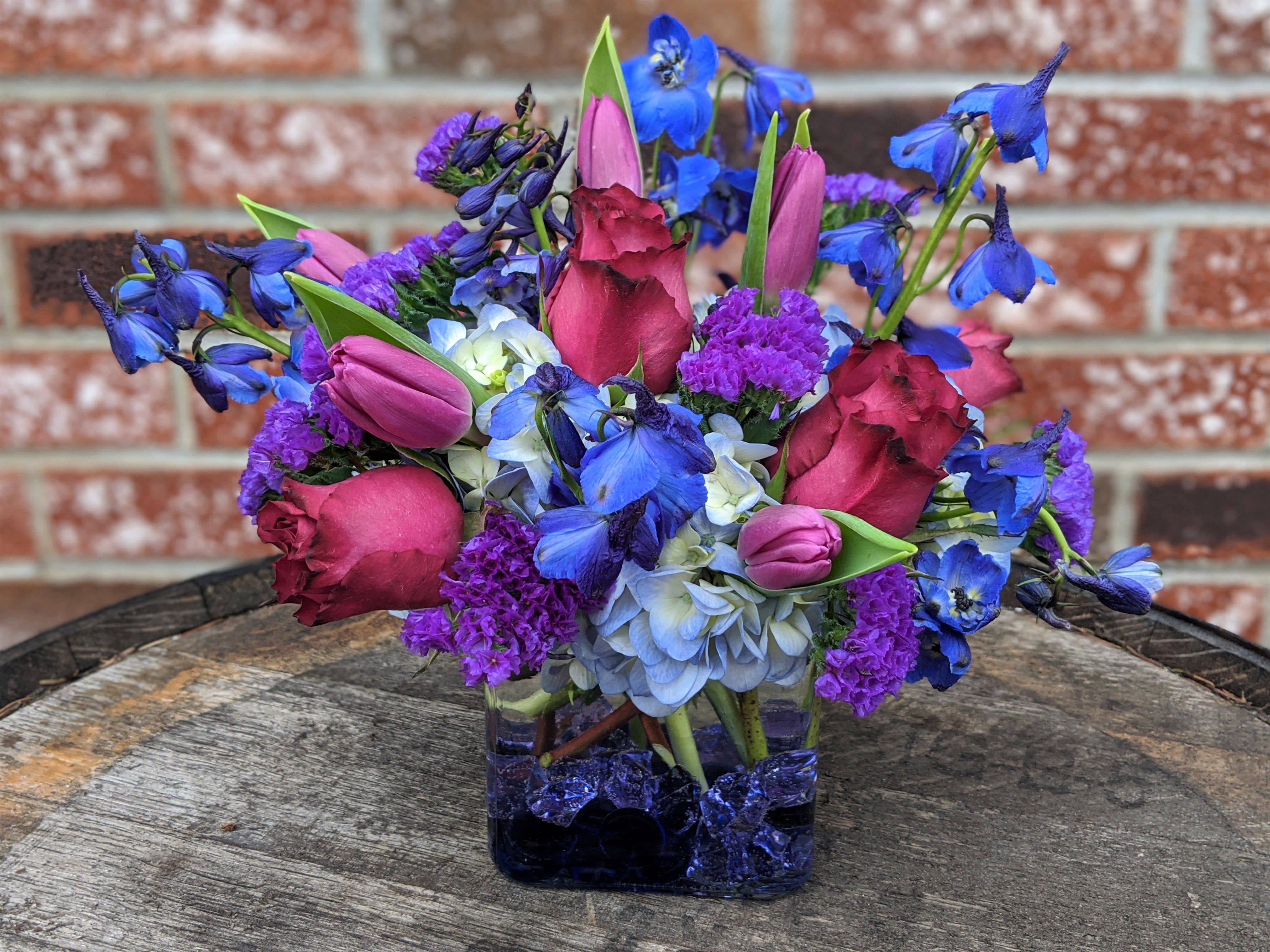 Blueberry Fields - Shown as Standard Blueberry roses, hydrangea, statice, tulips and delphinium comes arranged in a clear glass cube with accenting beads. *We custom design this arrangement and use the freshest flowers available the day of delivery. The arrangement in this picture is an example of the size and style and may not feature the exact product shown.*