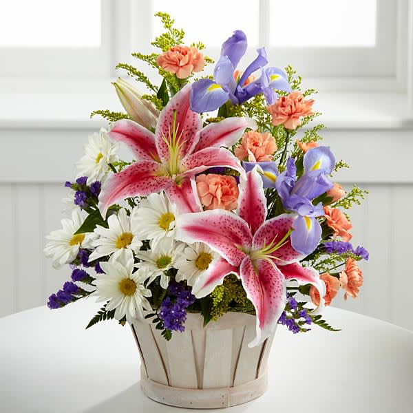 The Wondrous Nature Bouquet by FTD - targazer lilies stretch their fuchsia petals out amongst an arrangement of blue iris, white traditional daisies, orange mini carnations, purple statice and yellow solidago in a round whitewash handled basket.