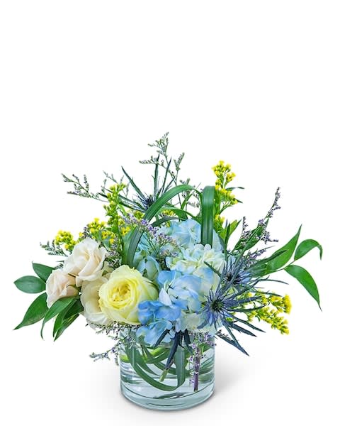 Wilde Blue - The soft blues and whites of this arrangement will add a touch of tranquility to any room. This peaceful bouquet combines blue hydrangea, white roses, blue eryngium, lily grass, solidago, Limonium, and Italian Ruscus in a clear glass vase. Wilde Blue is perfect for a sympathy gift.