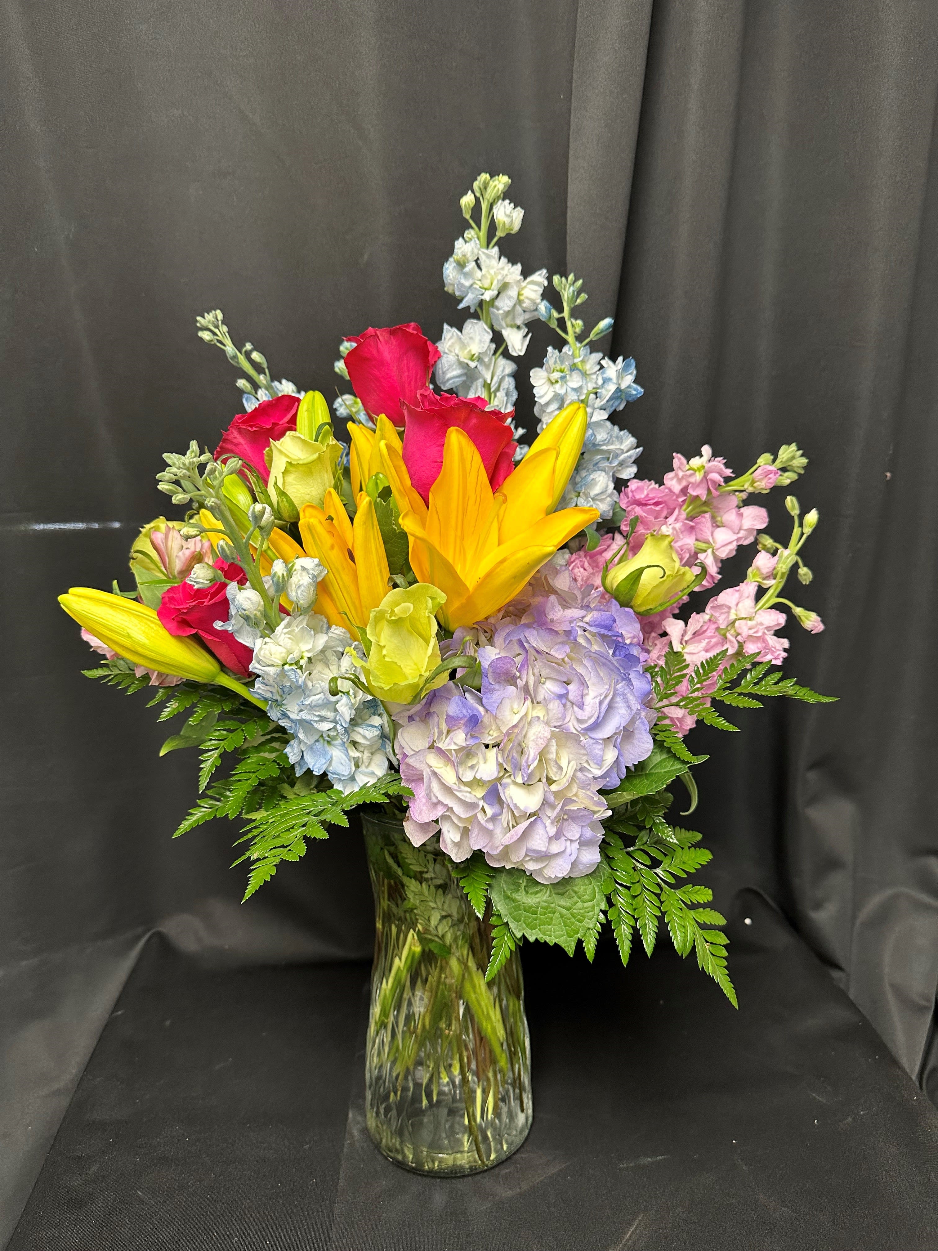Rainbow Brite -  Take a trip down memory lane to the vibrant days of the '90s with this exuberant arrangement of brightly colored blooms. Radiant yellow lilies stand tall amidst a riot of hot pink roses, pastel blue and pink stock, lush purple hydrangea, and cheerful yellow roses. This nostalgic bouquet is sure to evoke feelings of joy and nostalgia, bringing a burst of color and energy to any space.