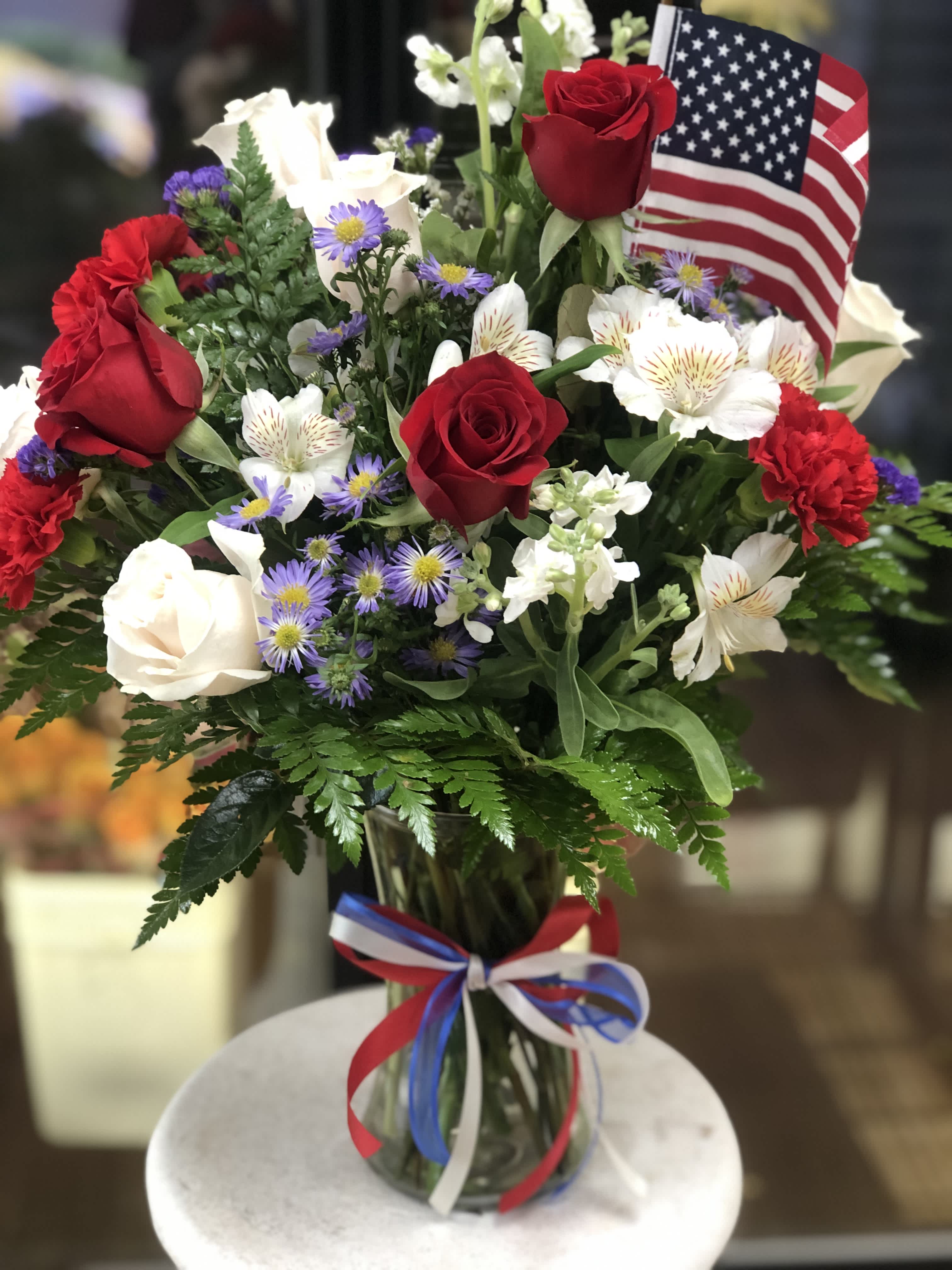 Liberty Blooms Liberty Blooms - The "Liberty Blooms" arrangement features a vibrant mix of red roses, carnations, and alstroemerias, complemented by montecasino, stock, and leather leaf greenery, creating a striking and patriotic display.