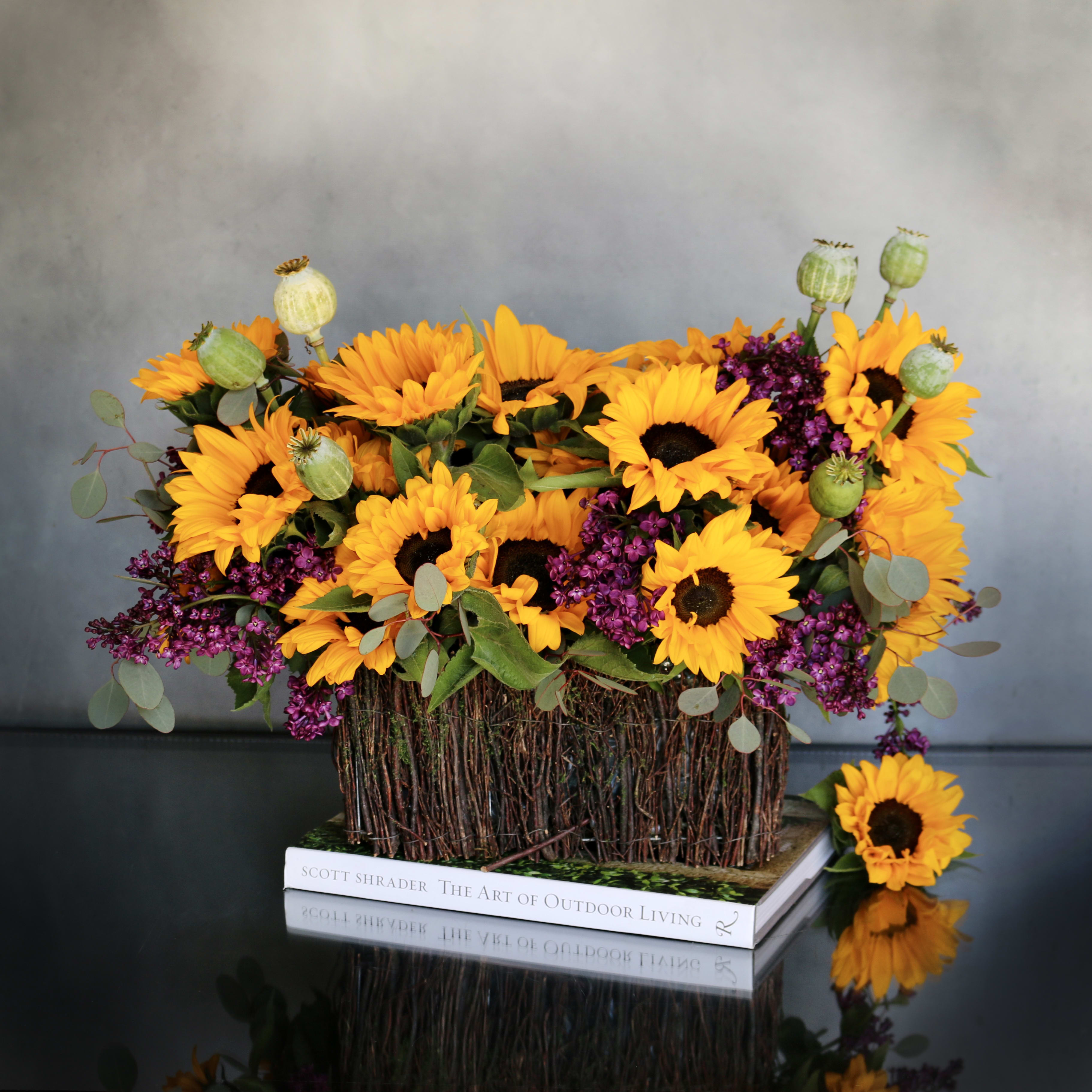 Sunny Smiles  - Container: 6 inches by 11 inches Arrangement: 13 inches by 20 inches This arrangement combines the bold beauty of sunflowers with the delicate allure of lilacs, creating a harmonious blend of colors and textures that will captivate the senses. Each bloom is carefully selected and artfully arranged to showcase its natural beauty, ensuring a stunning centerpiece that will brighten any space.  Whether you're celebrating a special occasion or simply want to bring a touch of nature indoors, this arrangement is sure to impress. Perfect for adorning a dining table, mantelpiece, or any other focal point in your home, it adds a touch of warmth and elegance to any setting.  Order now and let our gorgeous sunflowers and lilacs arrangement bring joy and beauty to your surroundings.