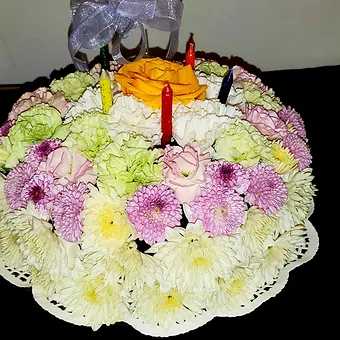 Floral Celebration cake - Our cheerful 9 inch birthday cake is set on a low cake plate and is made with a variety of flowers.  Made with white poms, purple buttons, and a rose in the center of the top of the cake.  Also comes with wax birthday candles.